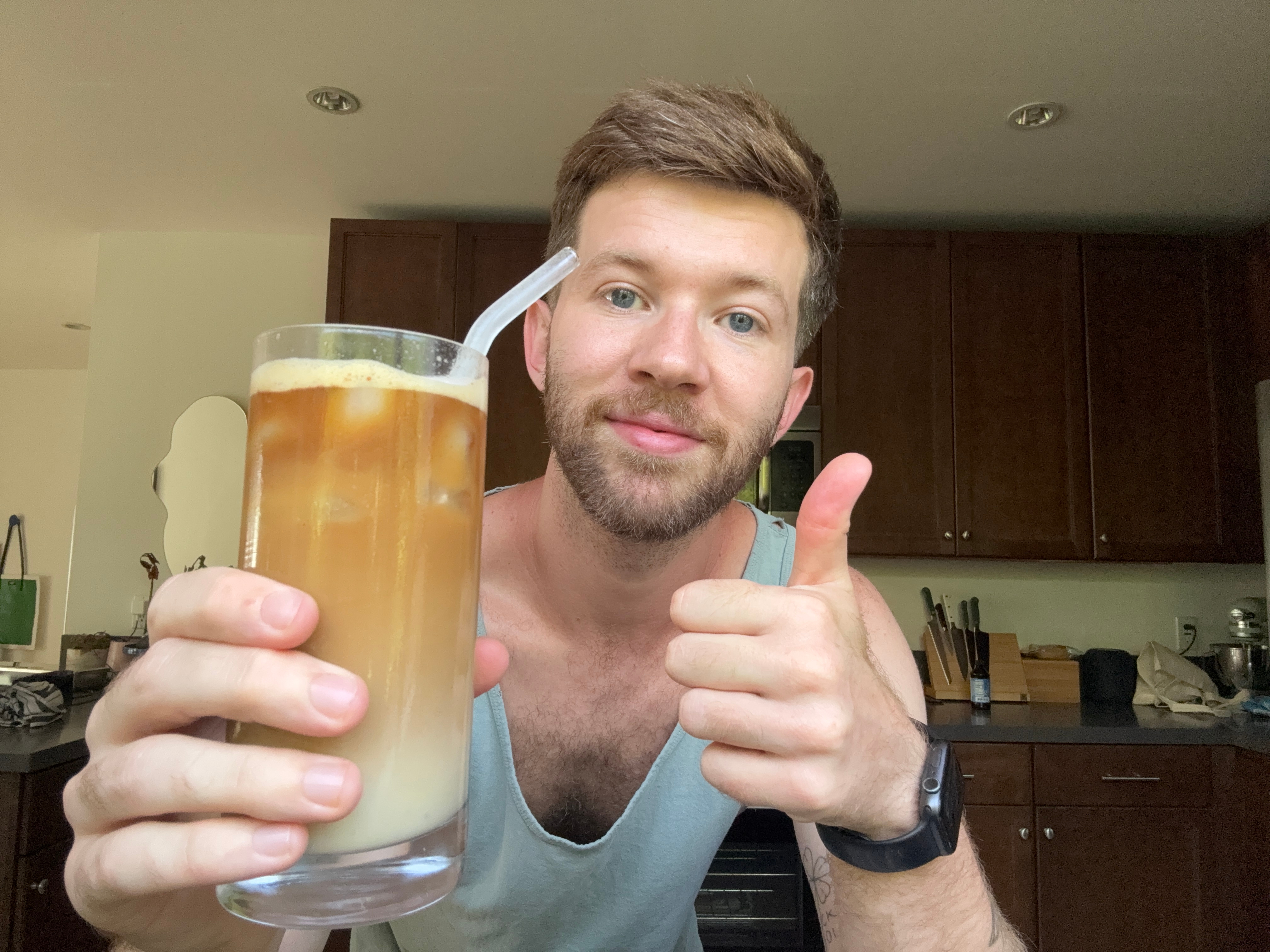 Person in a kitchen holding an iced coffee with a straw, giving a thumbs up, dressed in a casual tank top