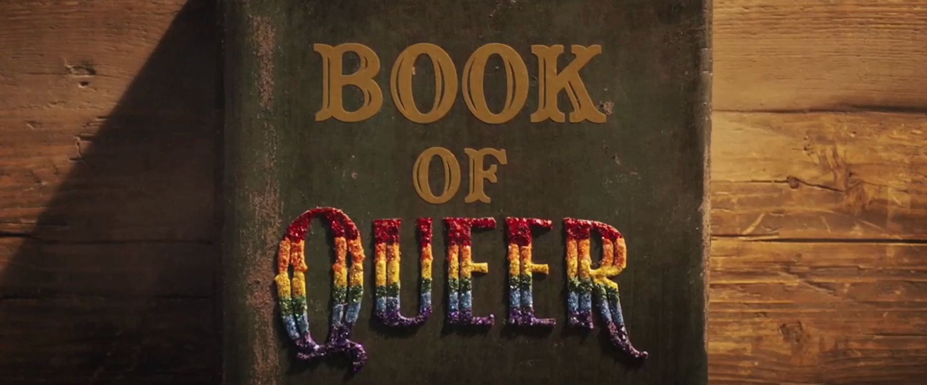 An old book titled "Book of Queer" with colorful, textured lettering on a wooden surface
