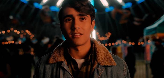 A young man with a thoughtful expression stands in an amusement park at night, wearing a denim jacket with lights blurred in the background