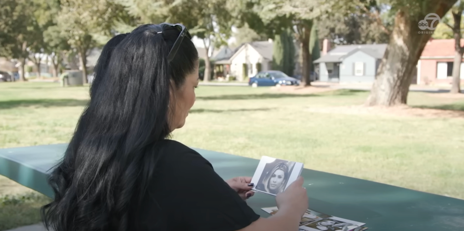 Person with long hair at a park bench, looking at black-and-white photos. Trees and houses are in the background