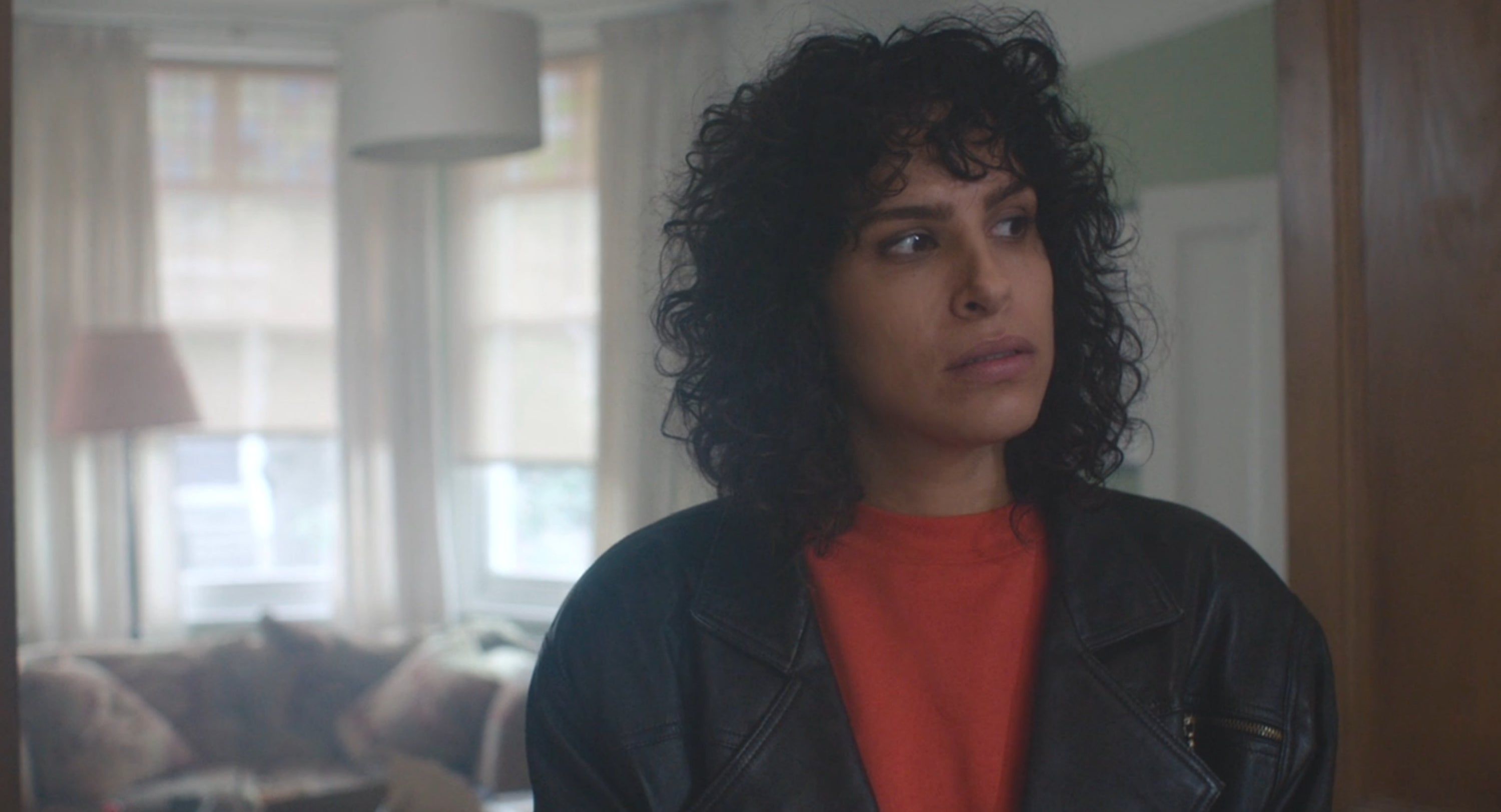 A person with curly hair looks contemplative in a living room, wearing a leather jacket over a red top