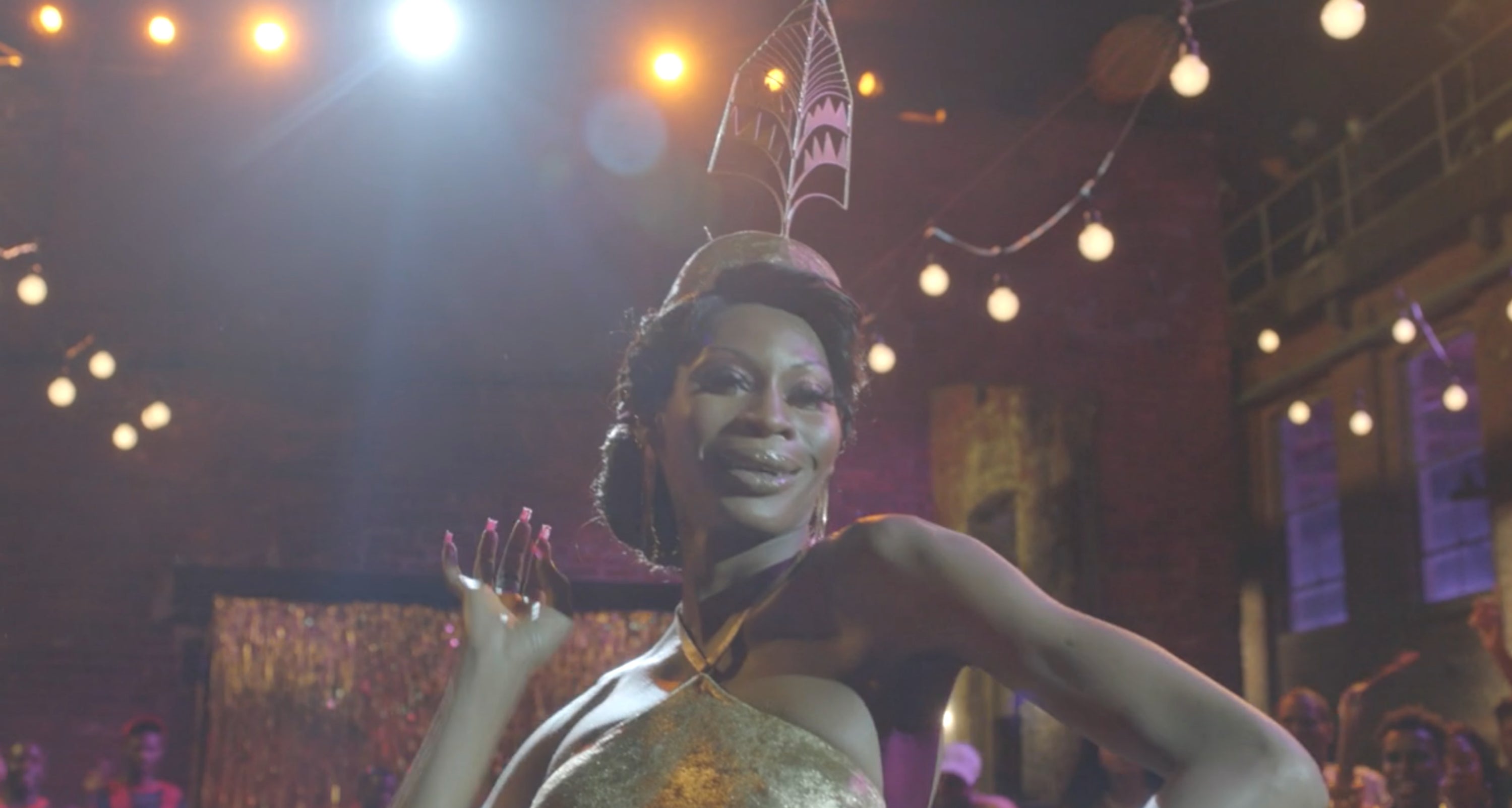 Person posing confidently at an event, wearing elegant, stylish attire and an artistic headpiece, with festive lights in the background