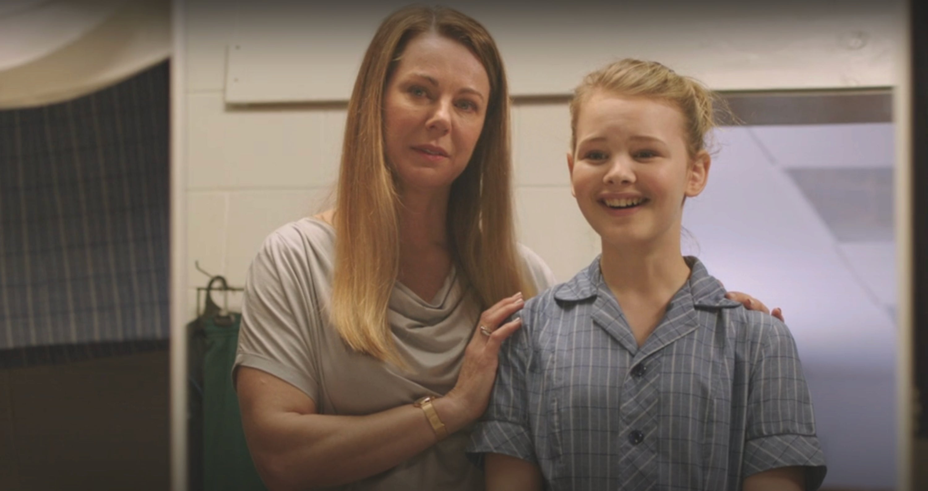 A woman standing with a young girl in a school uniform, both smiling in an indoor setting