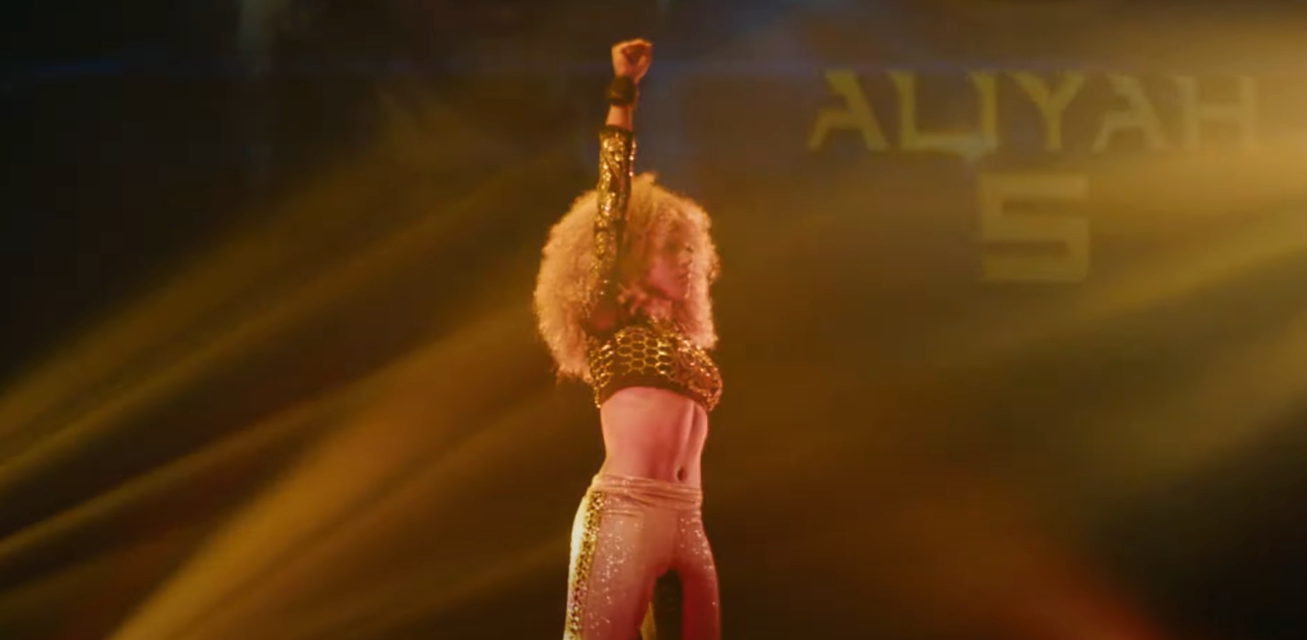 Performer with curly hair raises fist on stage in sequined top and pants, spotlight behind them. Stage background reads "ALIYAH 5."