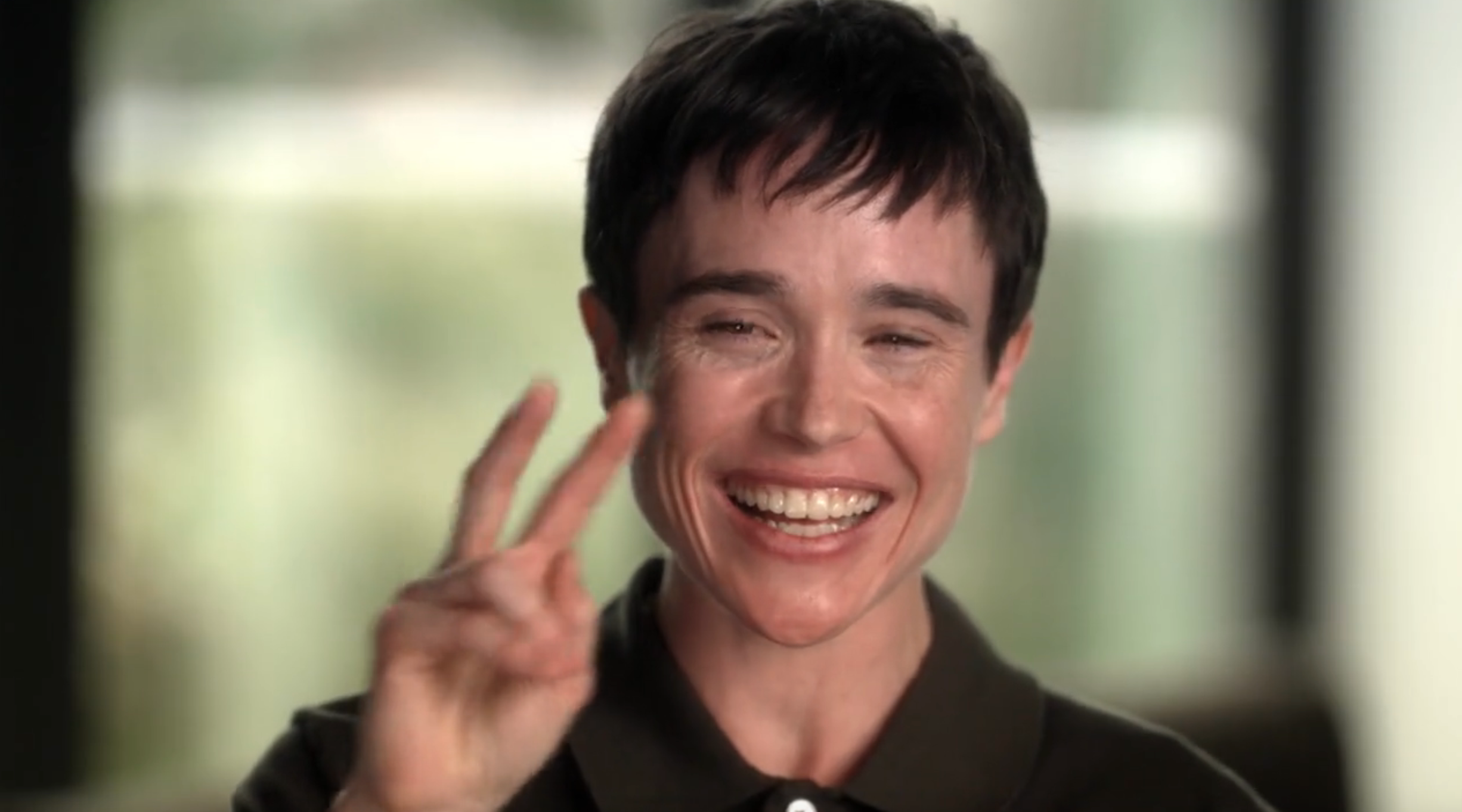 Person smiling, flashing a peace sign, wearing a short-sleeved polo shirt. Background is blurred and indoor setting