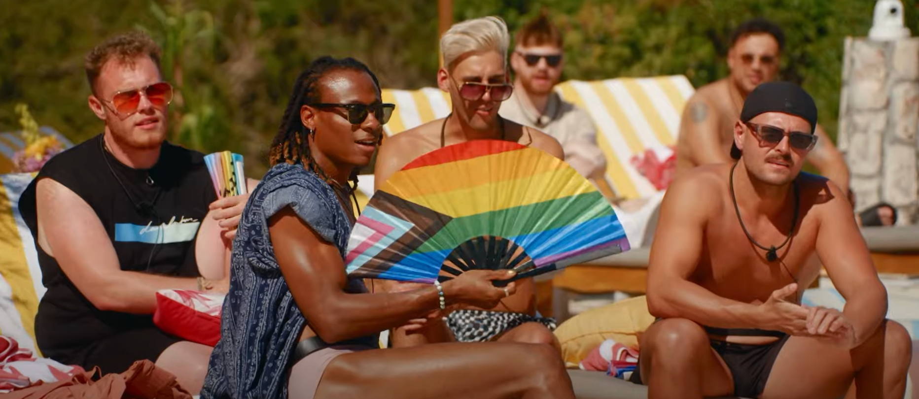 Group of people relaxing poolside, one holding a rainbow fan, wearing casual summer attire, including sunglasses and swimwear