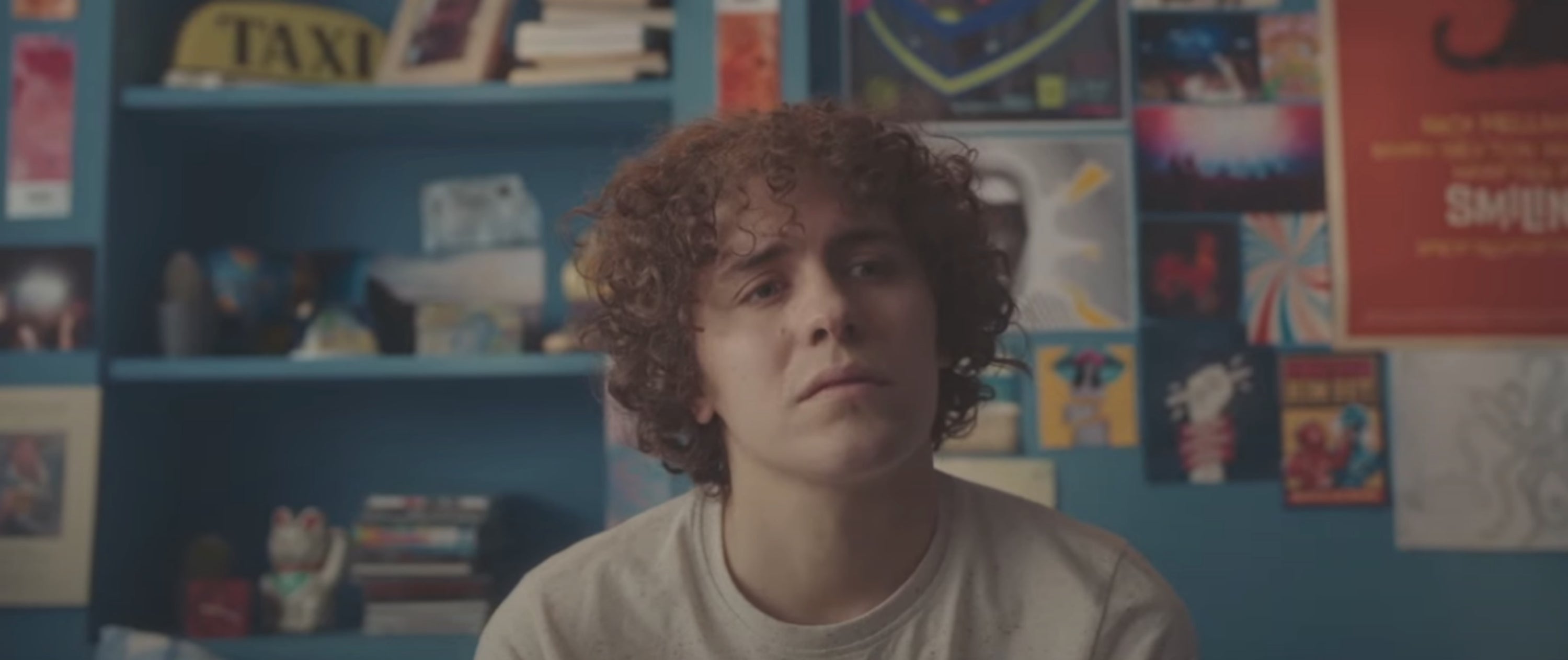 A person with curly hair sits in a room with posters and books on shelves, looking contemplative