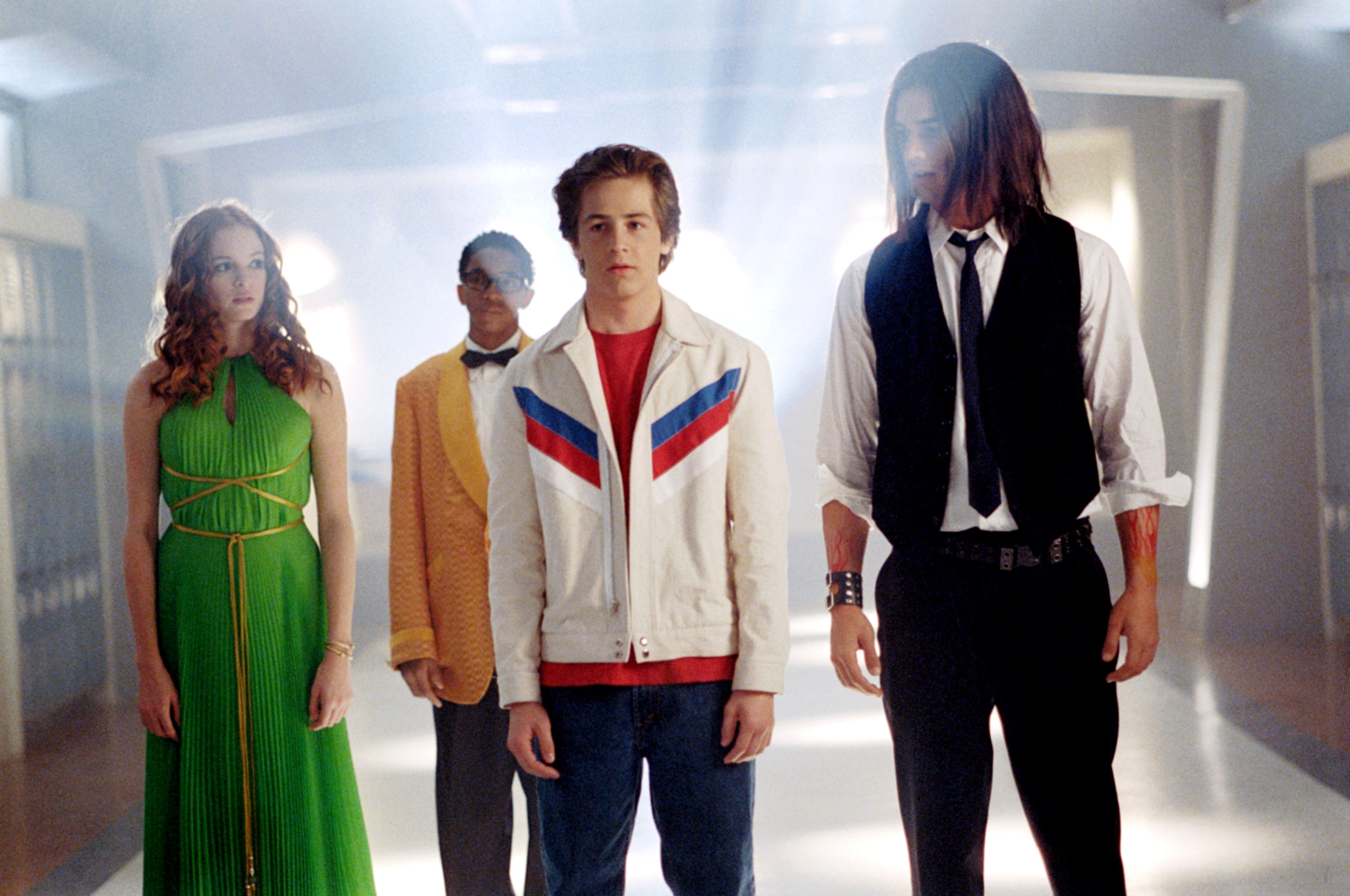 Four individuals stand in a school hallway: a person in a green dress, another in a yellow suit, one in a white jacket, and another in a vest and tie