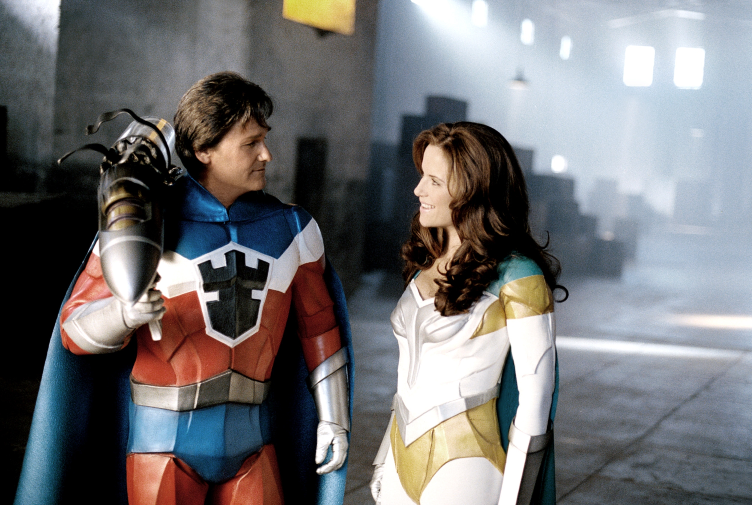 Two people in superhero costumes stand in a warehouse, smiling at each other. One holds a large device on their shoulder