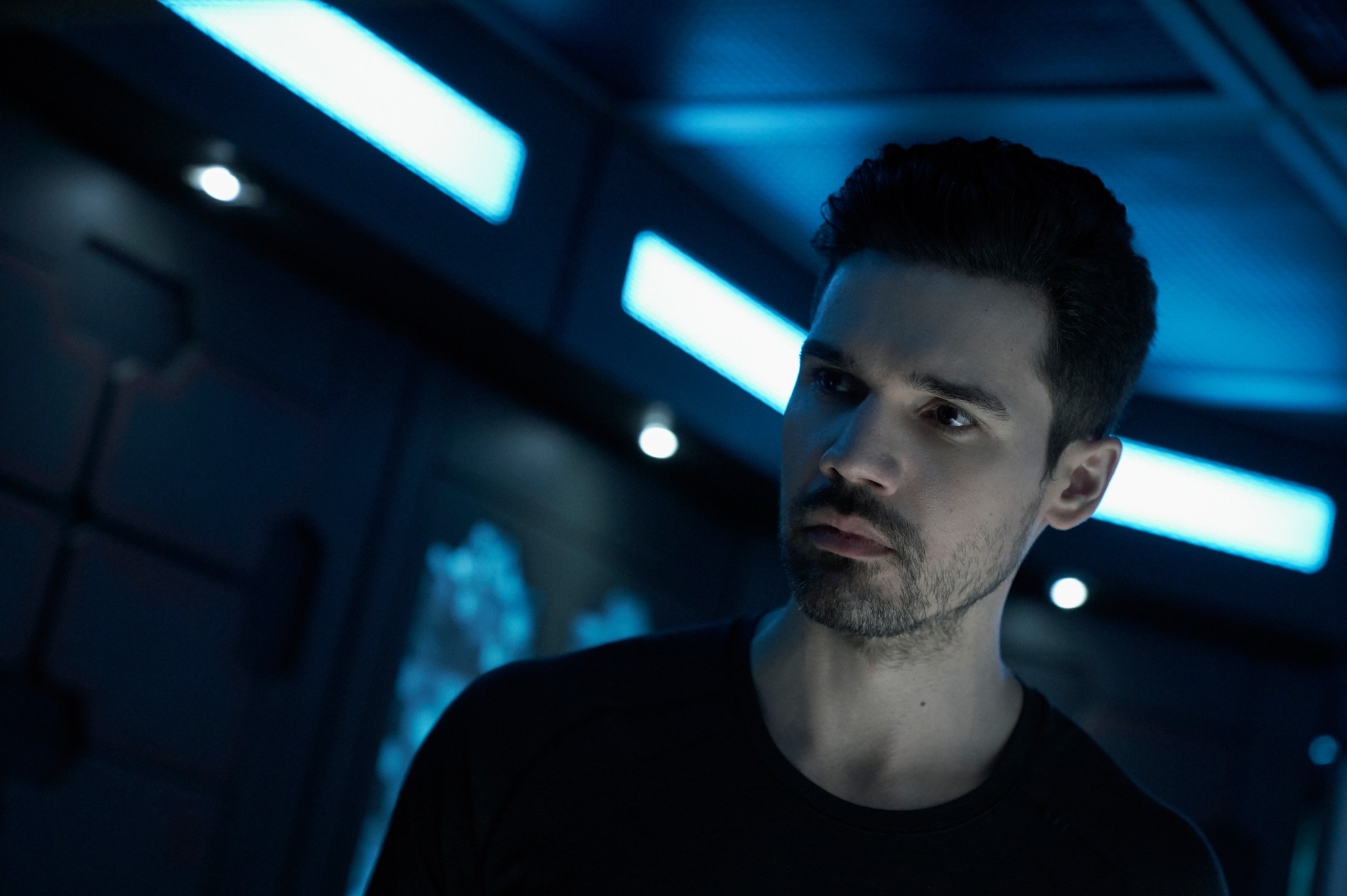 A person in a dimly lit sci-fi setting, looking serious, wearing a dark shirt