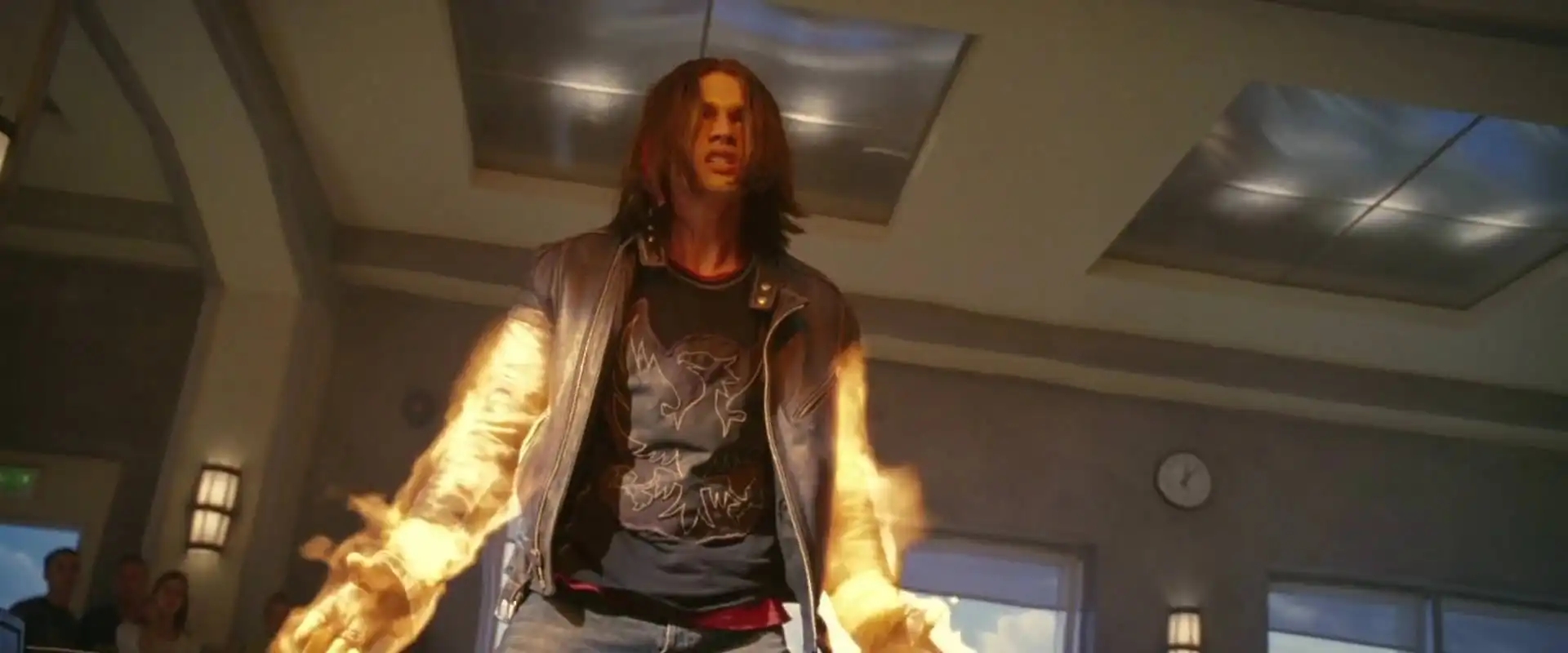 A person with long hair is inside a room, emitting flames from their arms, wearing a graphic t-shirt and jacket, conveying a dramatic moment