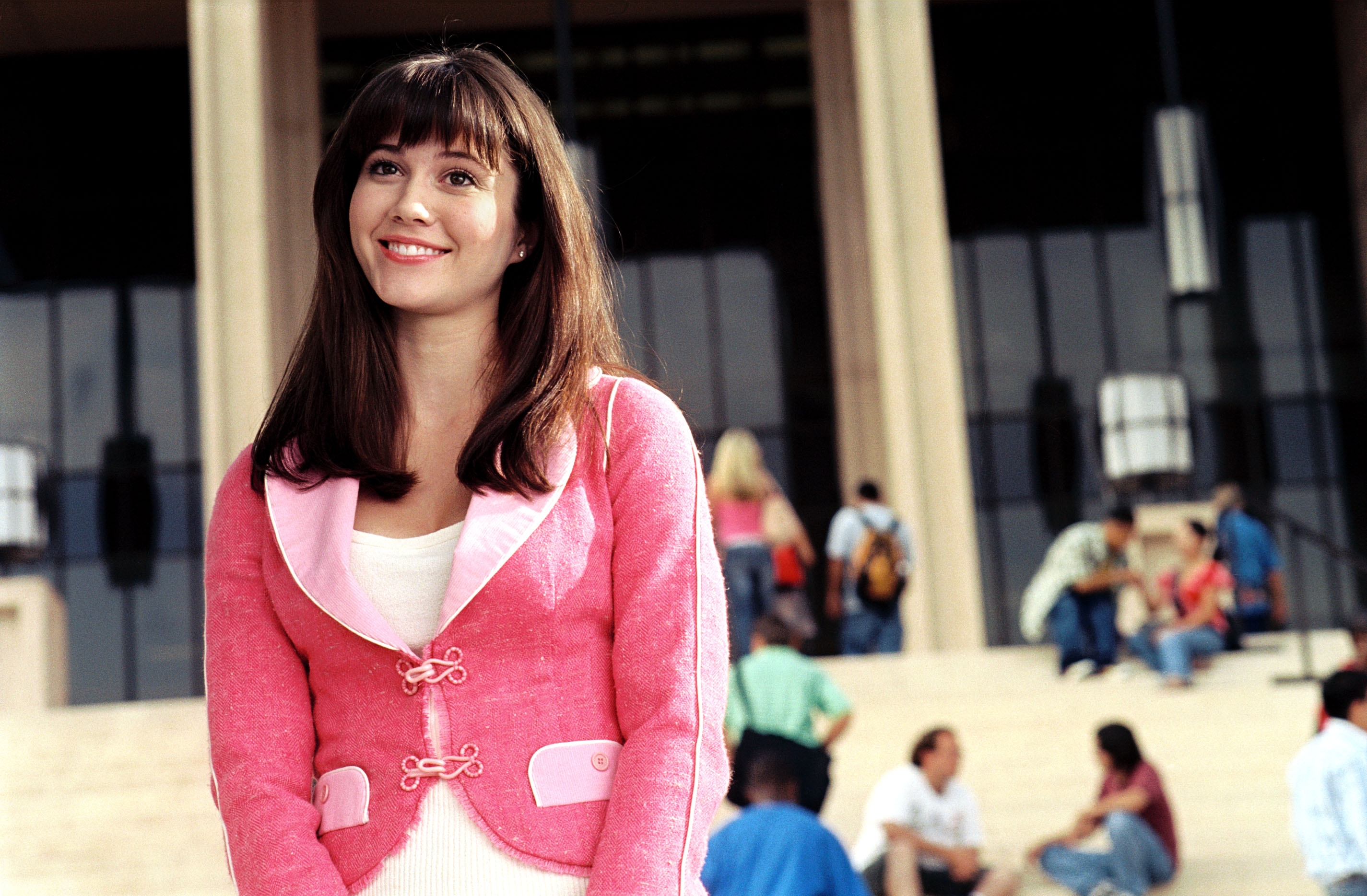 Person smiling, standing in front of a building with steps. They wear a stylish, pink jacket with a white top. People are seen in the background