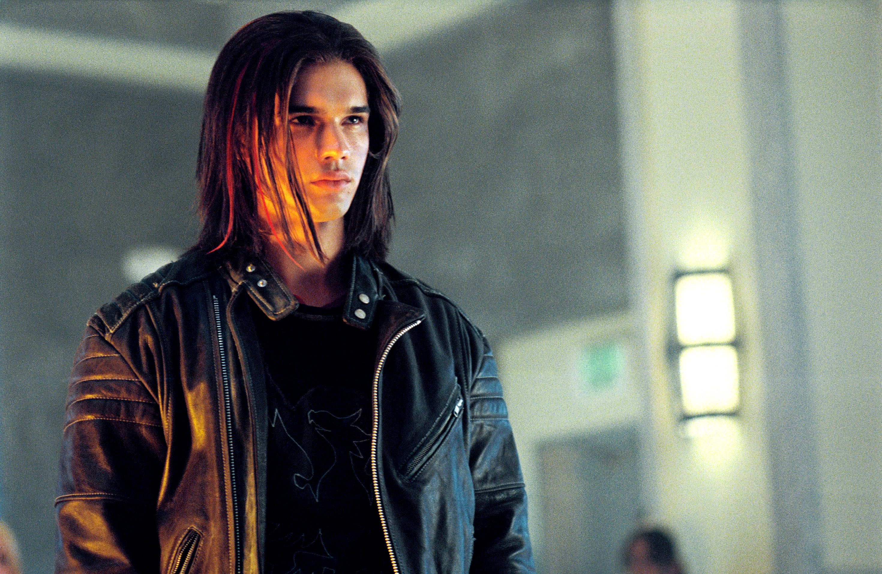 Person in a leather jacket with long hair, standing indoors; appears intense and focused