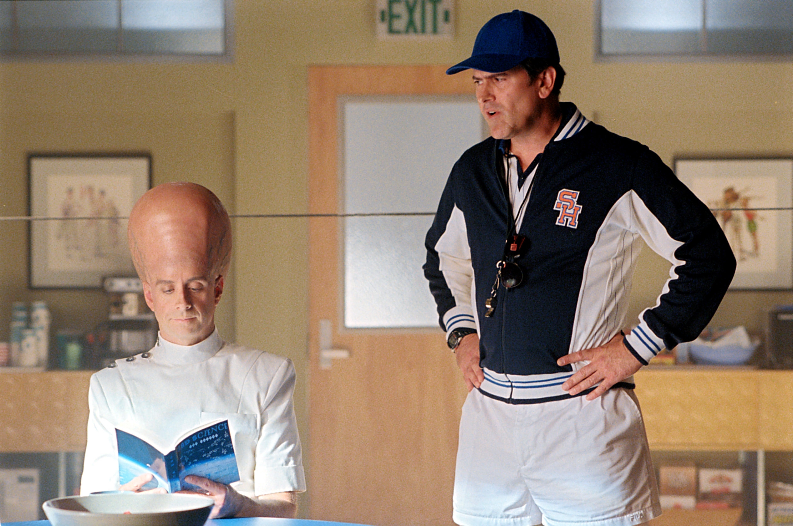 A character with an enlarged head reads a book, sitting beside a coach in a tracksuit and cap, in a comedic scene from a TV or movie set