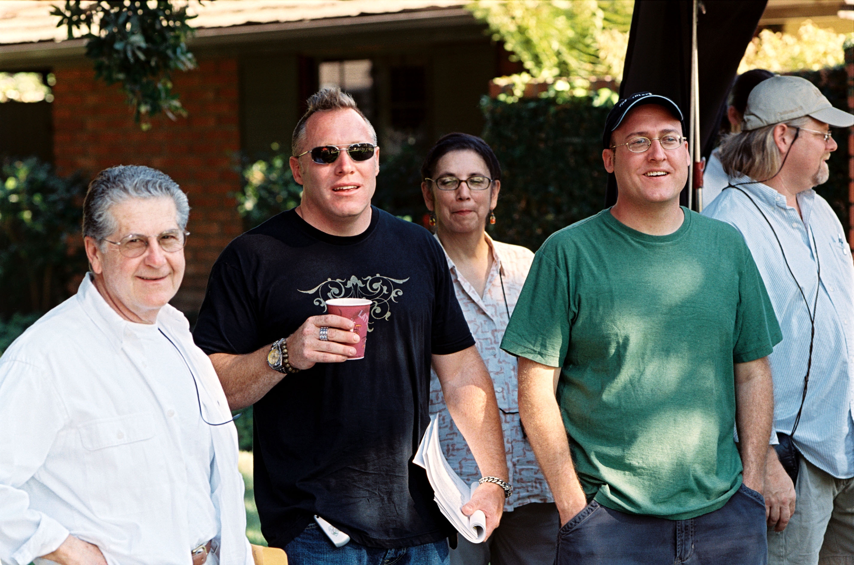 Group of five adults outdoors, casually dressed, some holding scripts or drinks, smiling. Trees and a building are in the background