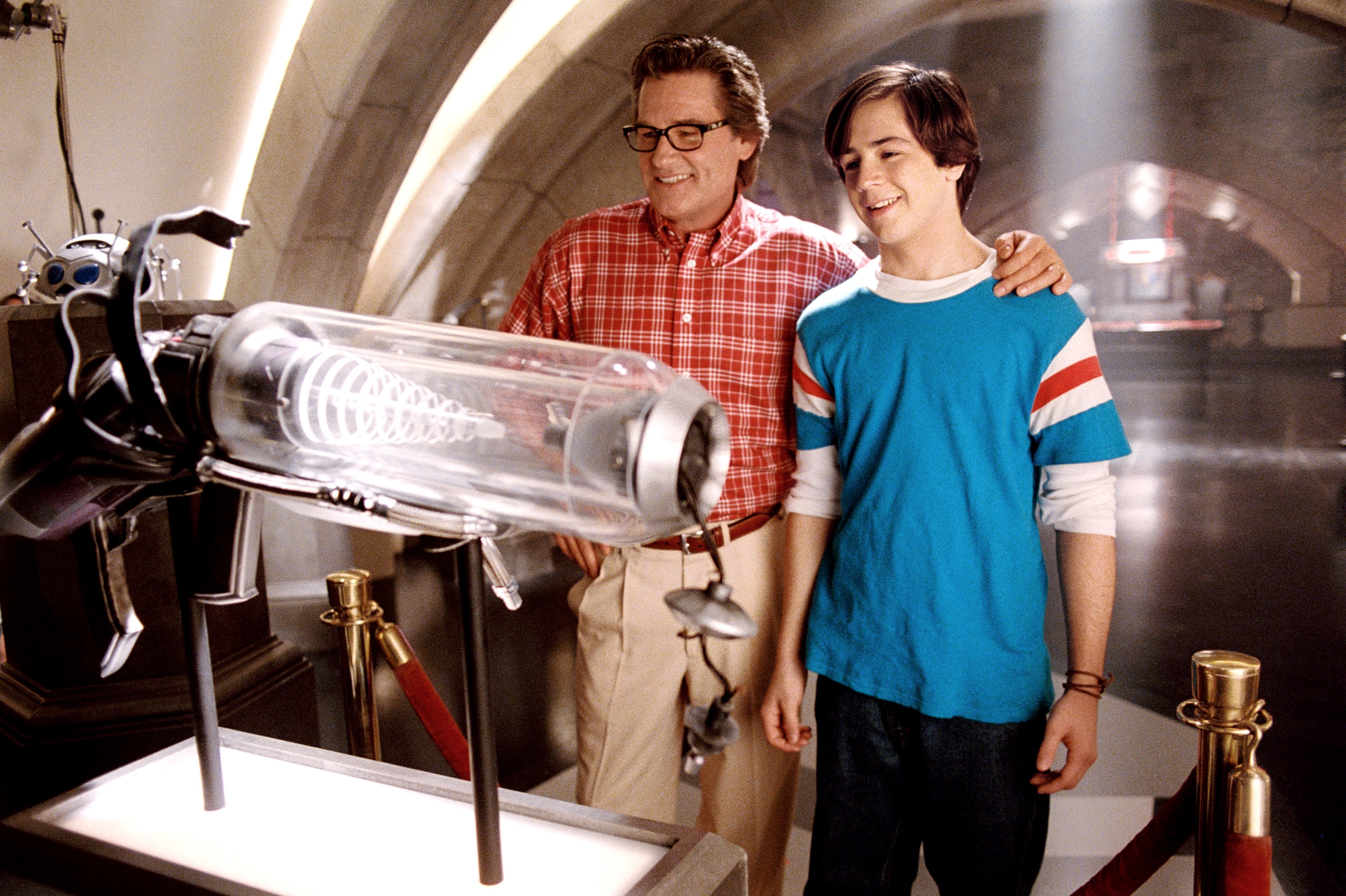 Two people in a lab setting, one wearing a plaid shirt and glasses, the other in a t-shirt with stripes, looking at a futuristic display