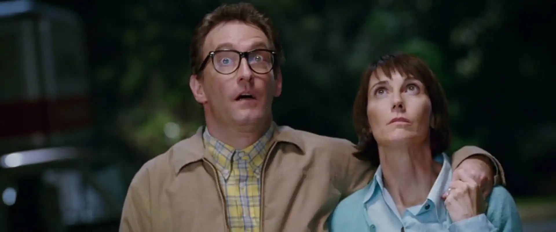 A man and woman look surprised and gaze upward, standing closely with his arm around her shoulders in an outdoor nighttime scene