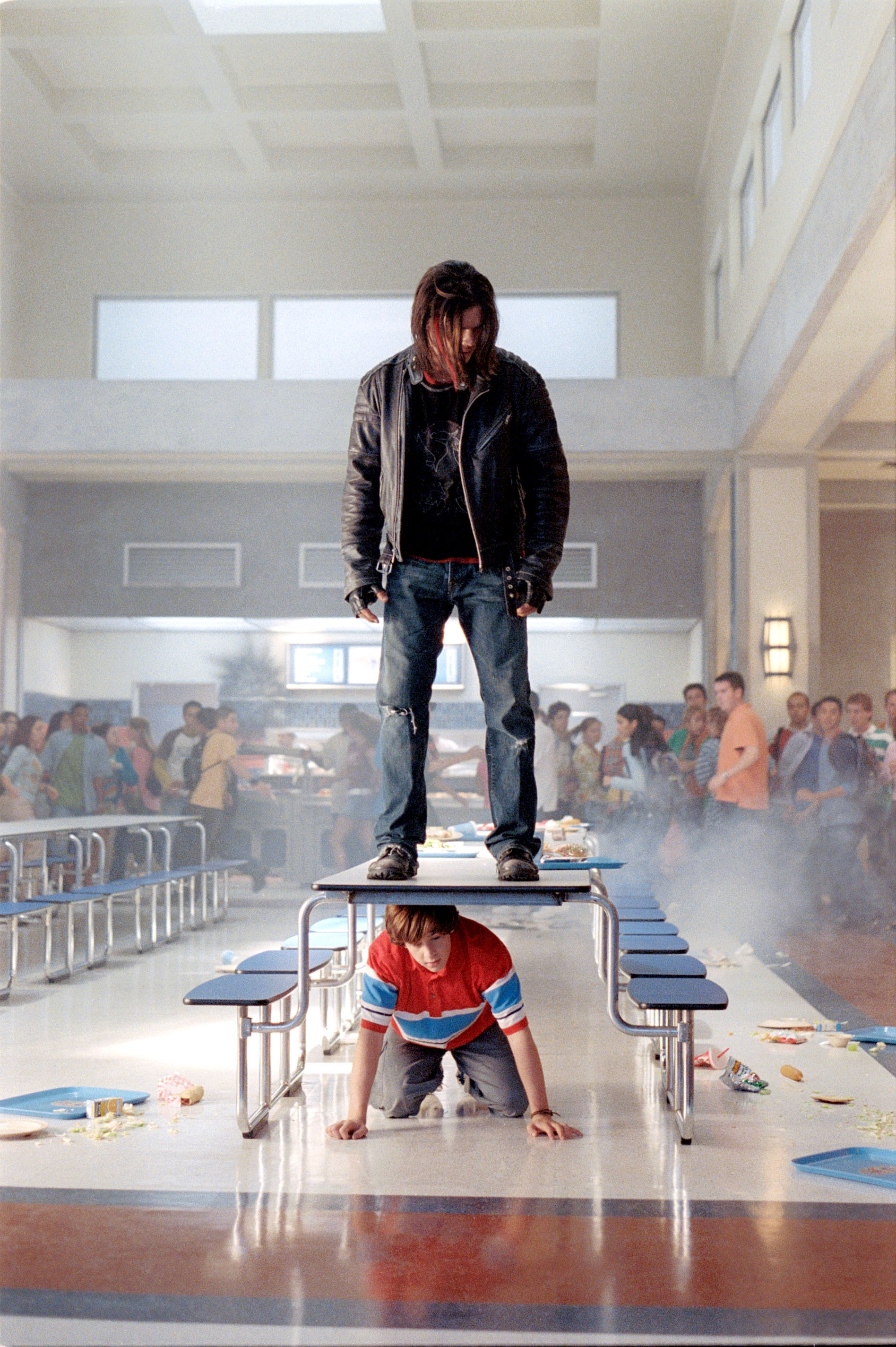 A person stands on a cafeteria table, looking down, while another crouches beneath it. Students stand around, observing the scene