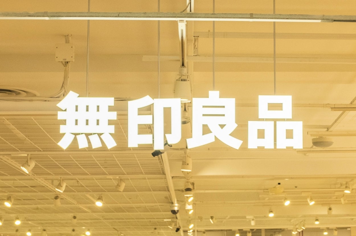 Shoppers browse inside a Muji store with various products on display. Large Japanese characters hang overhead