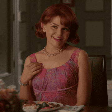 Woman in a vintage-style dress smiles and gestures, seated at a dining table set with food