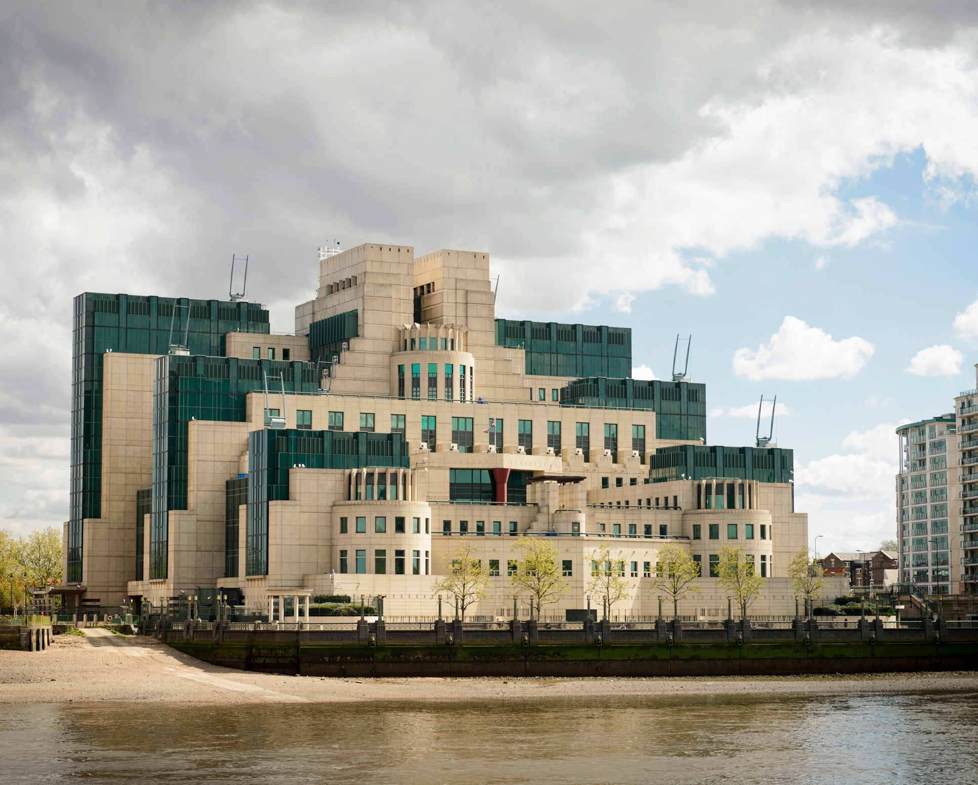 The image shows the MI6 headquarters, a modern building with a tiered design, located by the River Thames in London