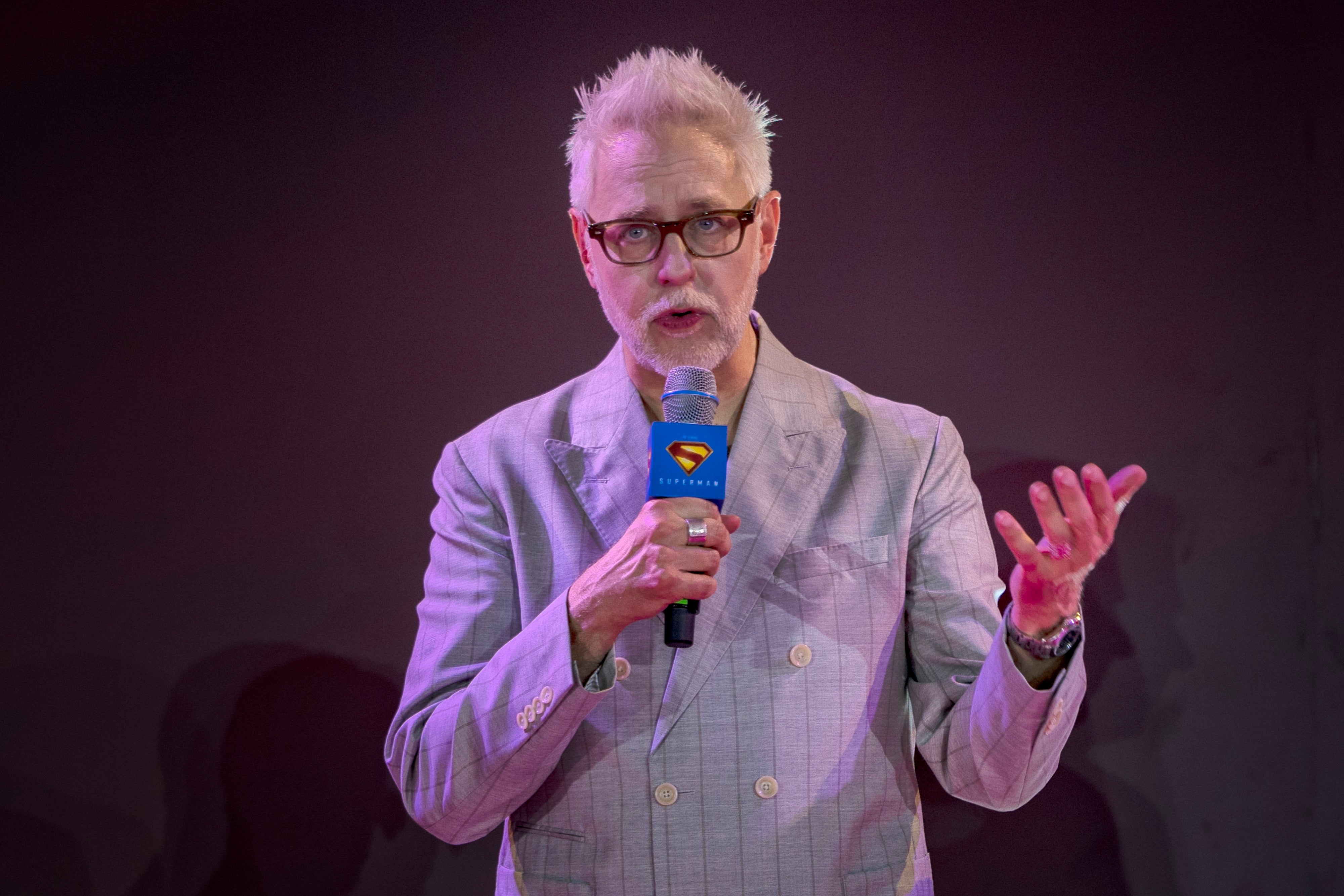 Man with glasses and styled hair speaks into a microphone, wearing a double-breasted suit jacket, gesturing with one hand