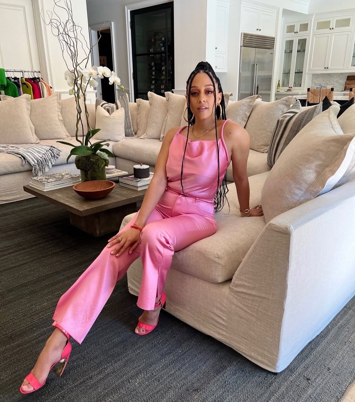 Person sitting on a sofa in a stylish, sleeveless pink outfit and matching shoes, posing in a modern living room