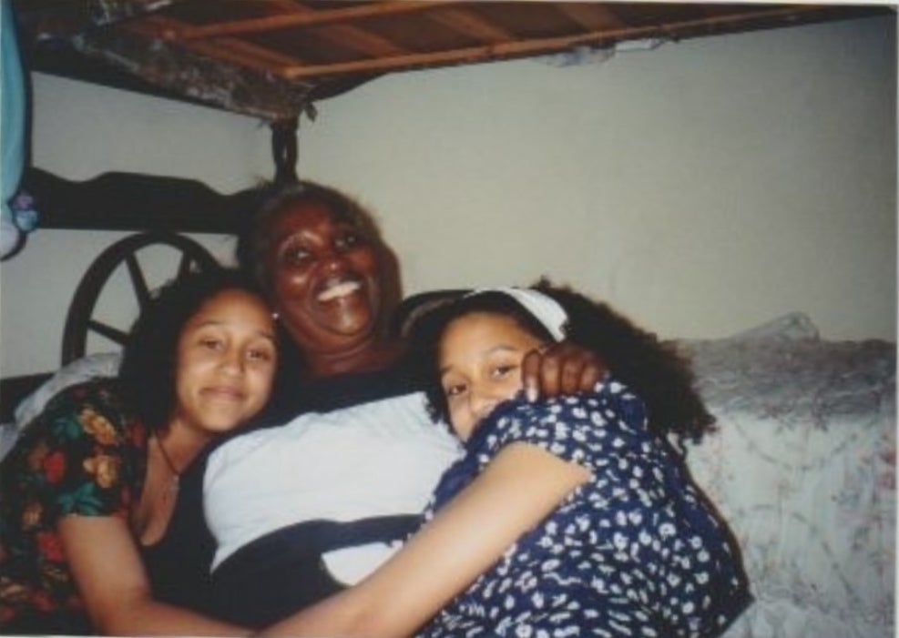 Two young women hug an older woman while sitting on a bed, smiling for the camera. The setting appears to be informal and cozy