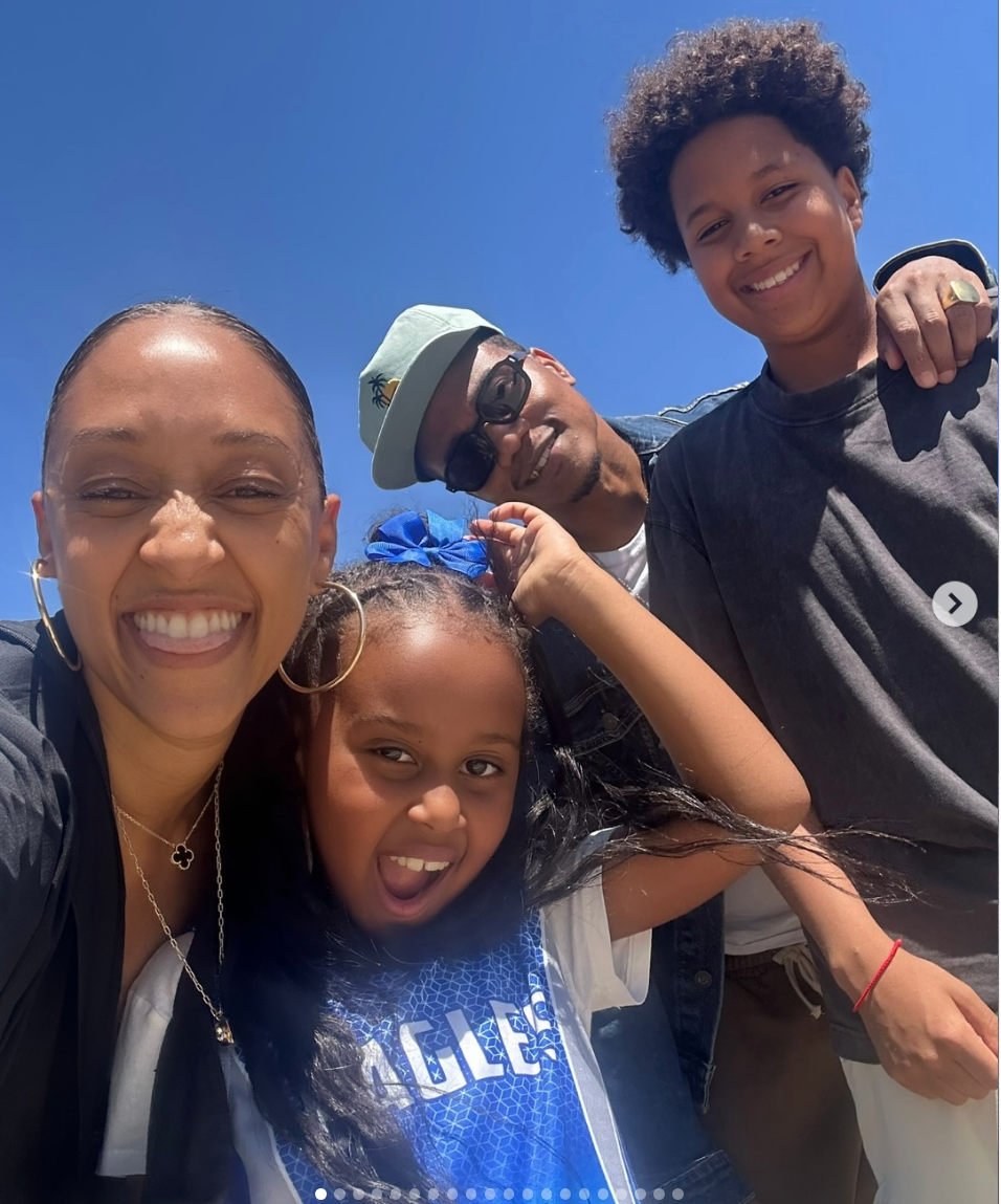 A smiling group of four people poses outdoors. The front child wears a sports jersey and bow, while the adults and older child wear casual attire