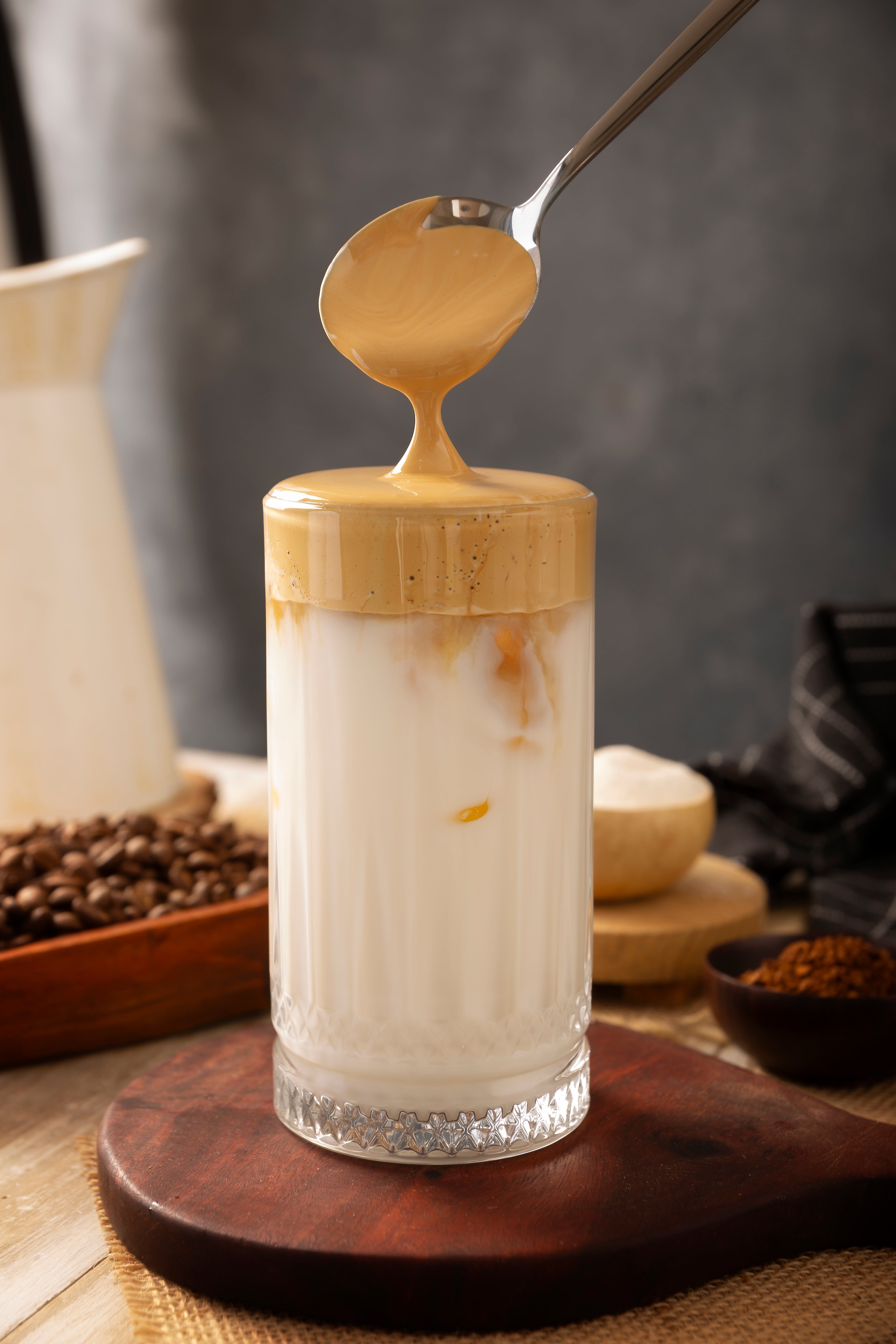 A spoon drizzles whipped coffee over a glass of iced milk, surrounded by coffee beans and powder in a cozy setting