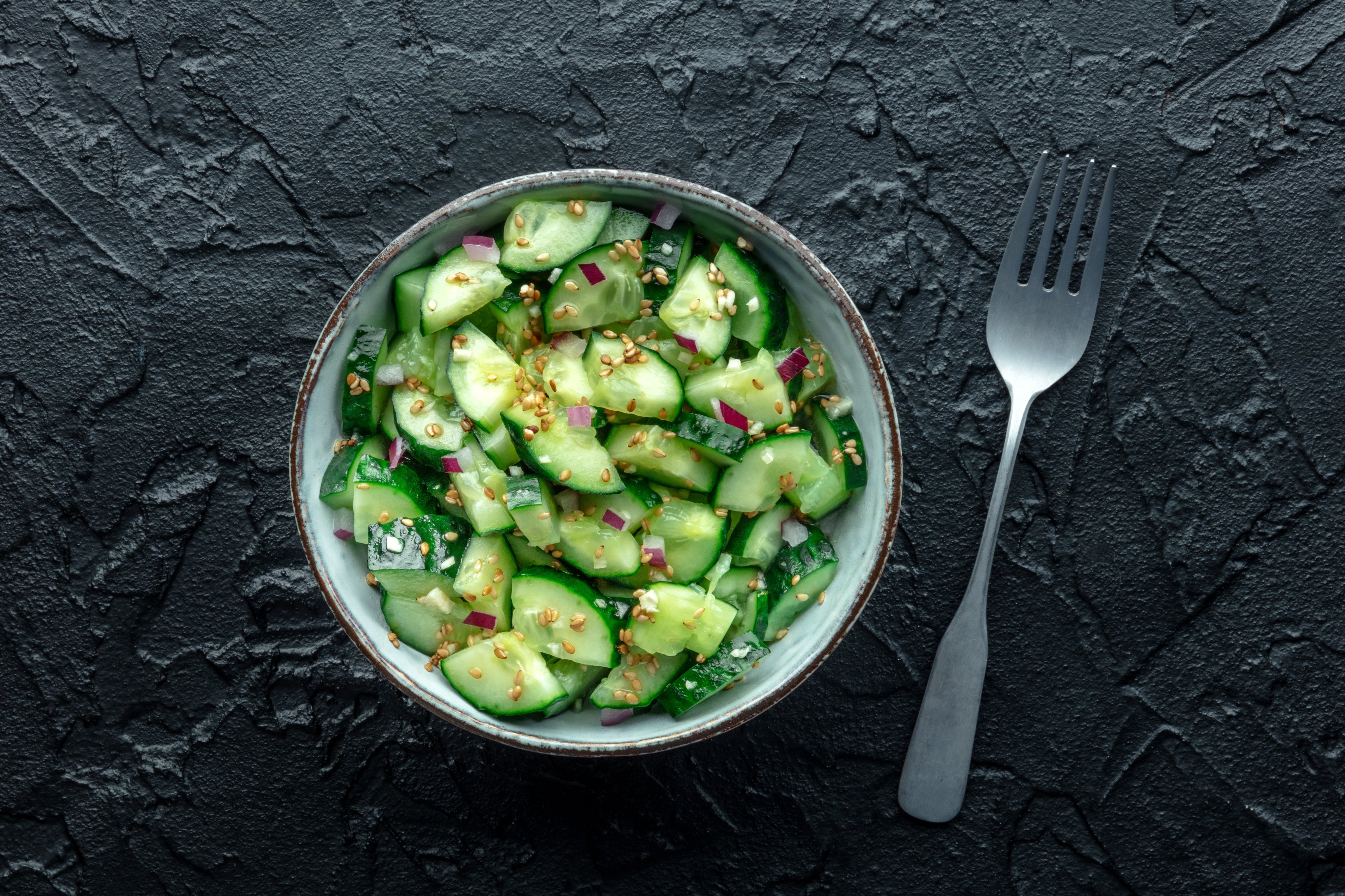 Bowl of cucumber salad with diced onions and seasoning, on a textured dark surface, next to a fork