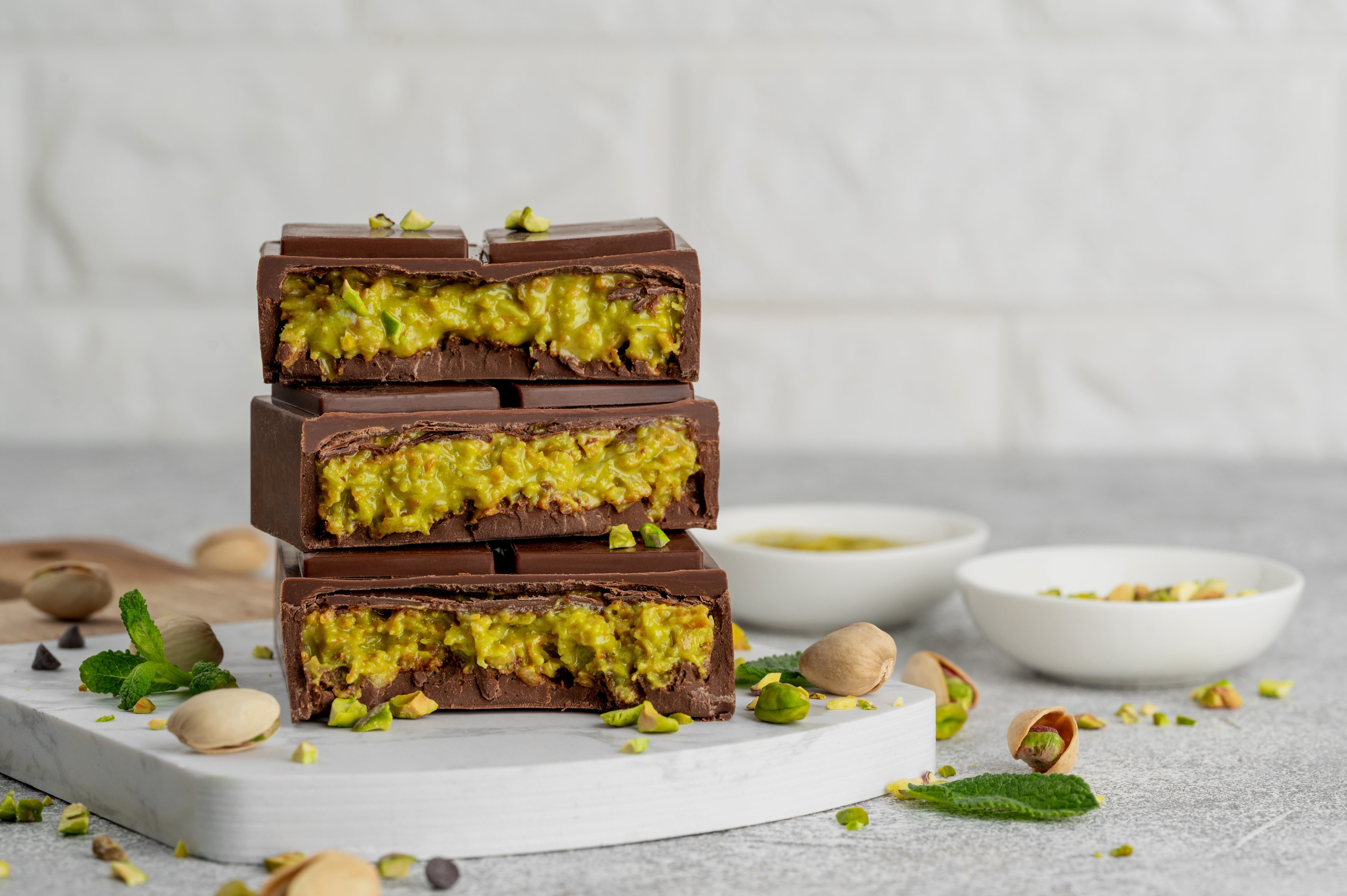 Three stacked chocolate bars filled with a pistachio mixture, placed on a countertop with scattered pistachios and mint leaves