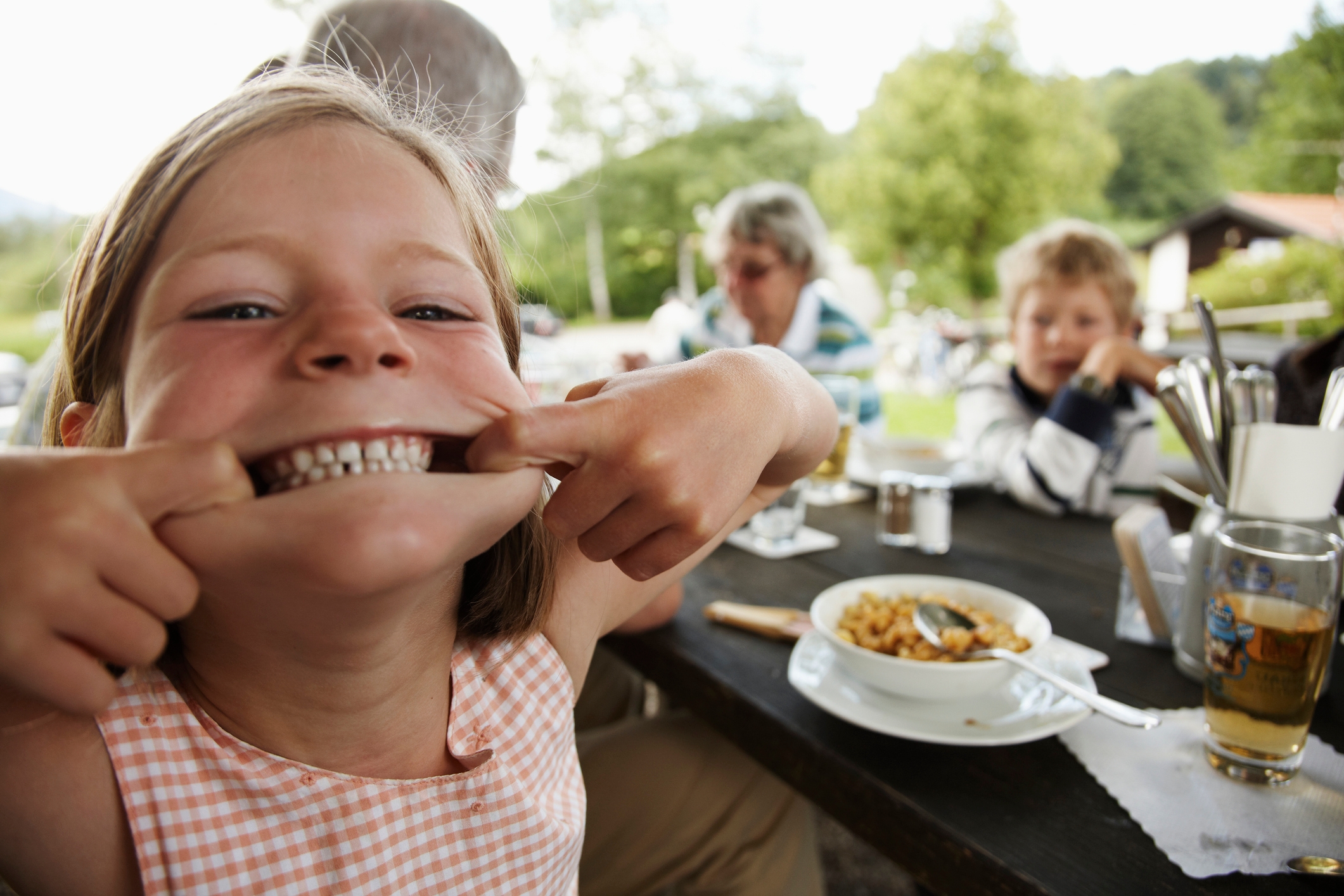 Barnet strekker lekent munn, smiler ved utendørsbordet med spisestuen. Andre mennesker og mat er i bakgrunnen, og skaper en uformell, gledelig scene