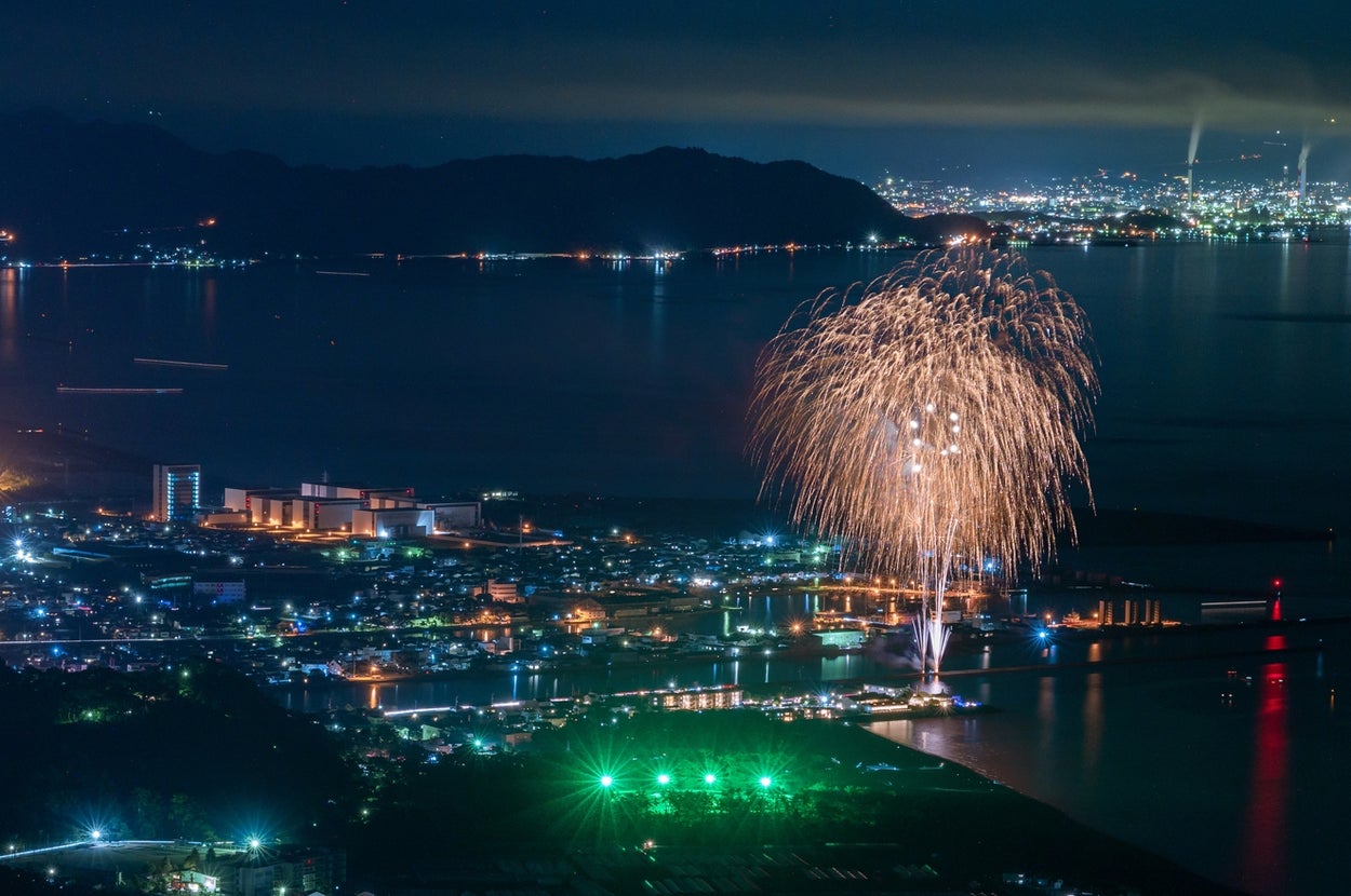 都市の夜景と湖のそばで打ち上げられる花火。