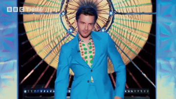 Person in a bright blue suit and colorful shirt dances confidently on stage with a large wheel backdrop