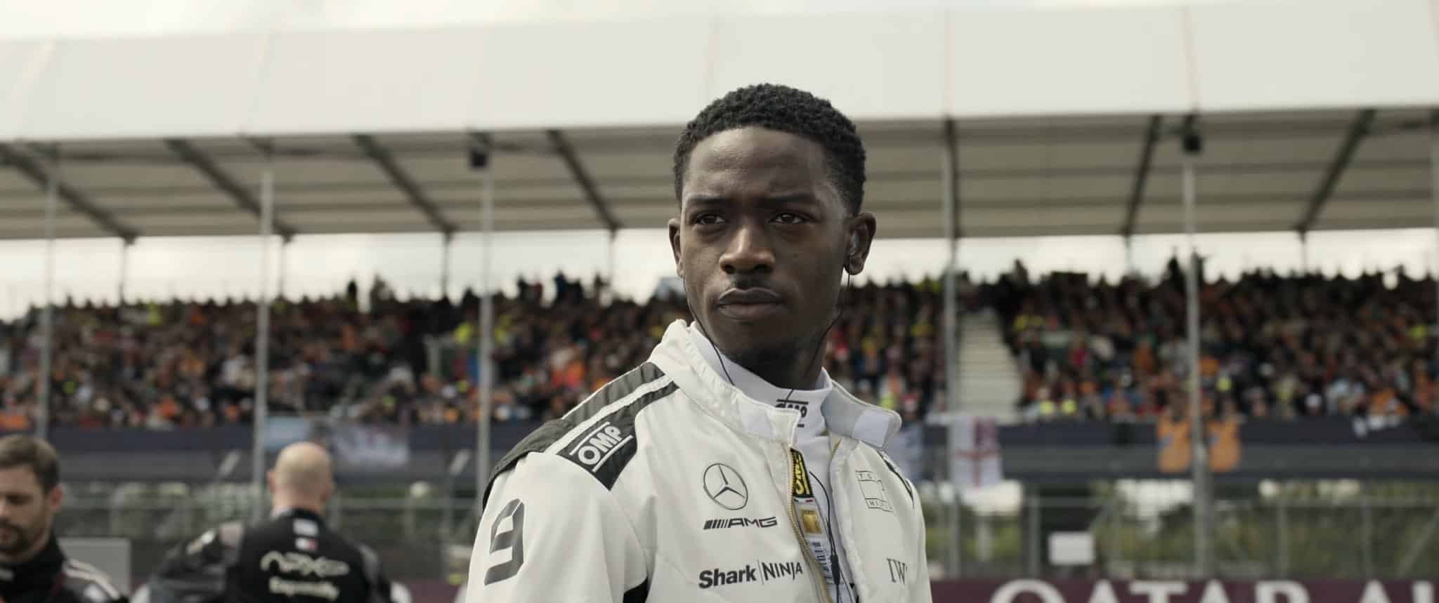 Person in a racing suit stands on a racetrack, crowd in the background