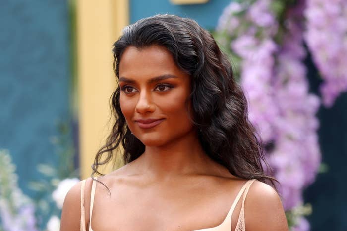 A person with long wavy hair on a red carpet, wearing an elegant sleeveless gown, poses in front of floral decorations