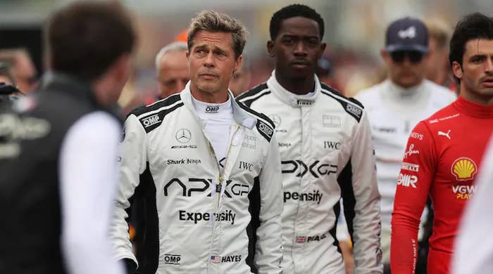 Two people in racing suits stand among others at an event, looking focused. The suits feature sponsor logos and badges