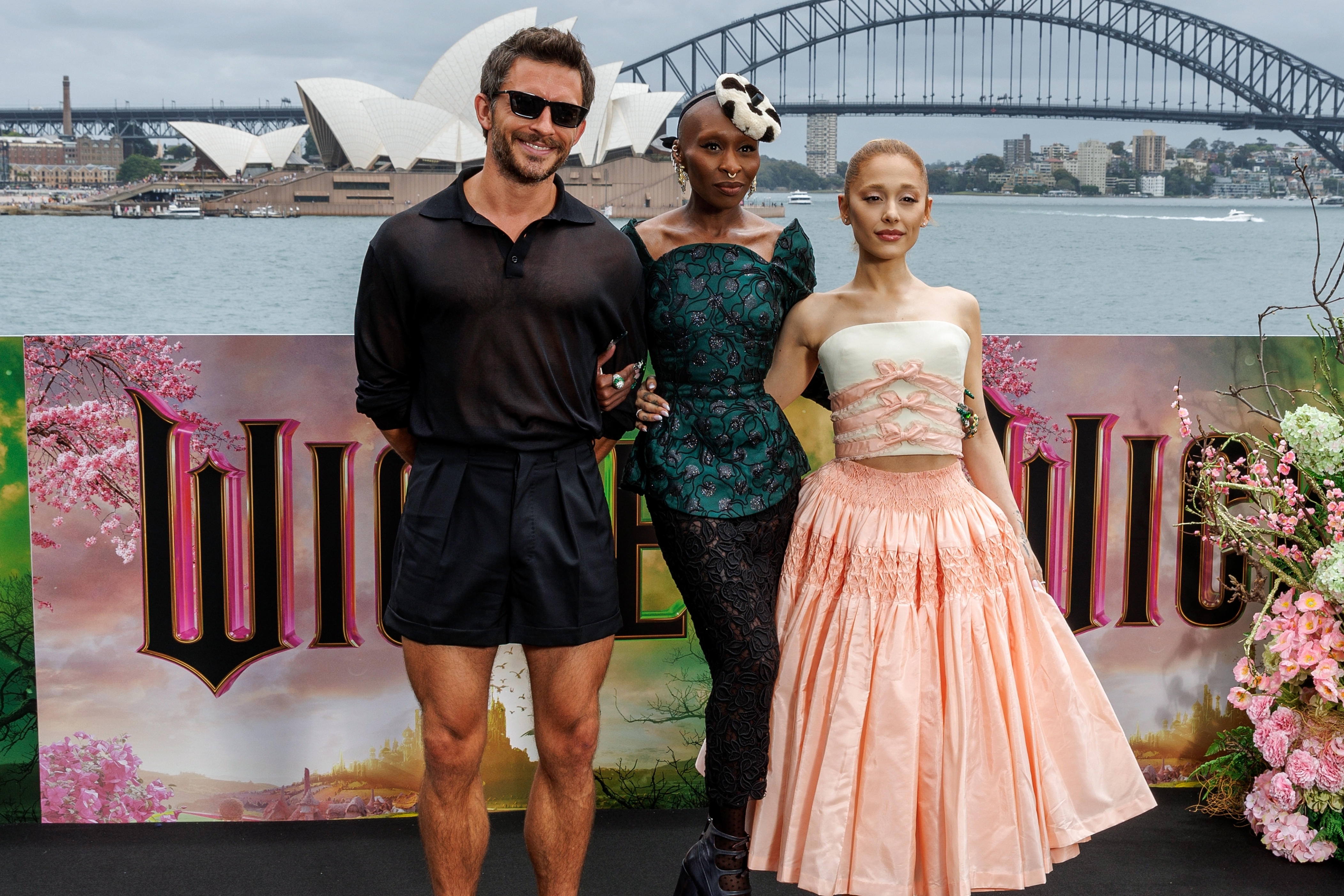 Three people pose at a "Wicked" event by Sydney Harbour. One person wears a casual outfit, another wears a structured top with patterned pants, and the third wears a strapless dress