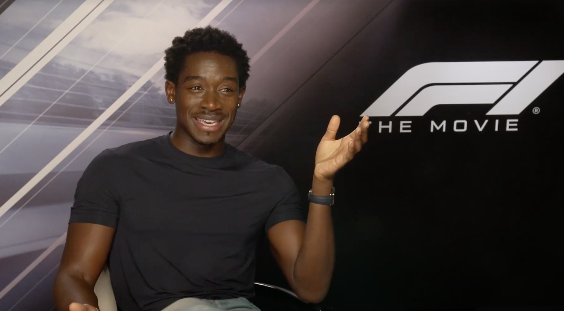 Person seated, gesturing enthusiastically during an interview, with "F1 The Movie" logo in the background. Casual attire with a t-shirt