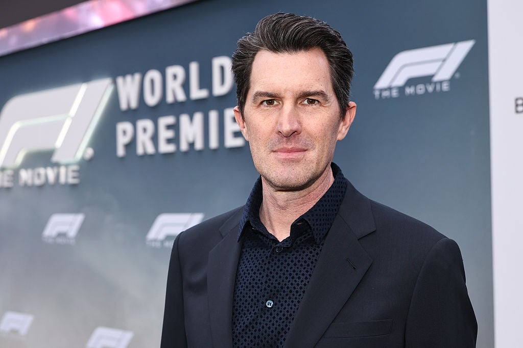 A person in a suit jacket and patterned shirt at a film premiere event. The backdrop displays "World Premiere" with a movie logo