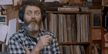 Man with headphones and a plaid shirt nods and raises eyebrows, standing in front of a shelf filled with records
