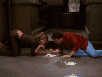 Two people are on the floor eating cheesecake with forks, humorously and intently focused on their task