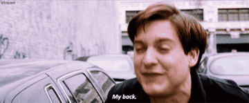 Person bends over dramatically in discomfort, exclaiming, &quot;My back,&quot; while standing next to parked cars on a street