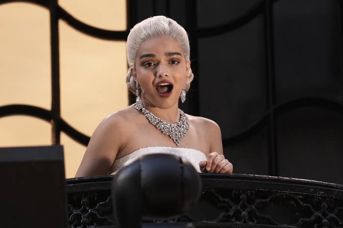 Rachel Zegler with elegant hairstyle and ornate jewelry sings on a balcony, wearing a strapless dress
