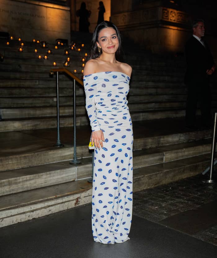 Rachel Zegler in an off-the-shoulder, patterned gown stands outside on a stone staircase, smiling softly