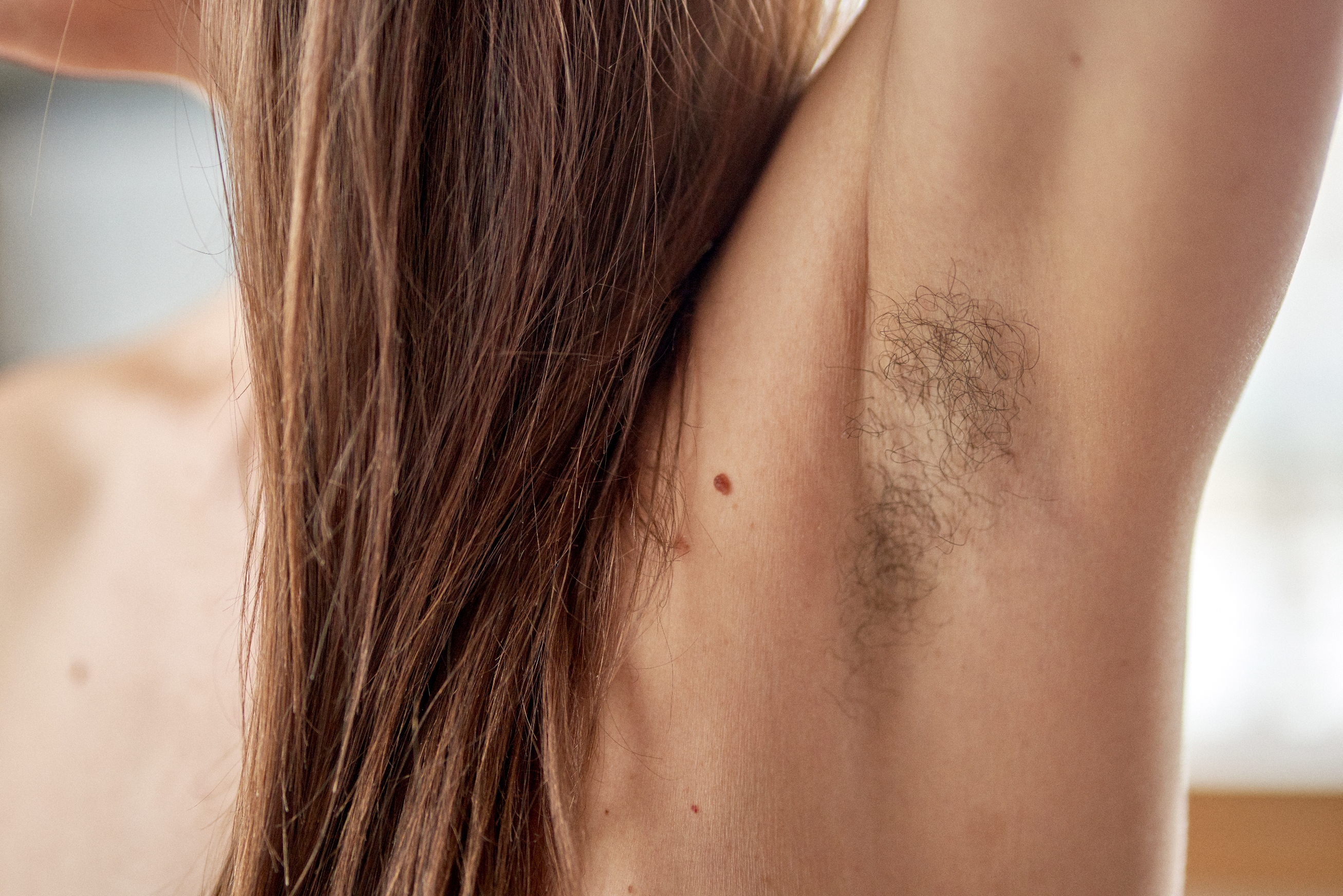Person showing natural underarm hair with a small mole on the side. Long hair falls down their back