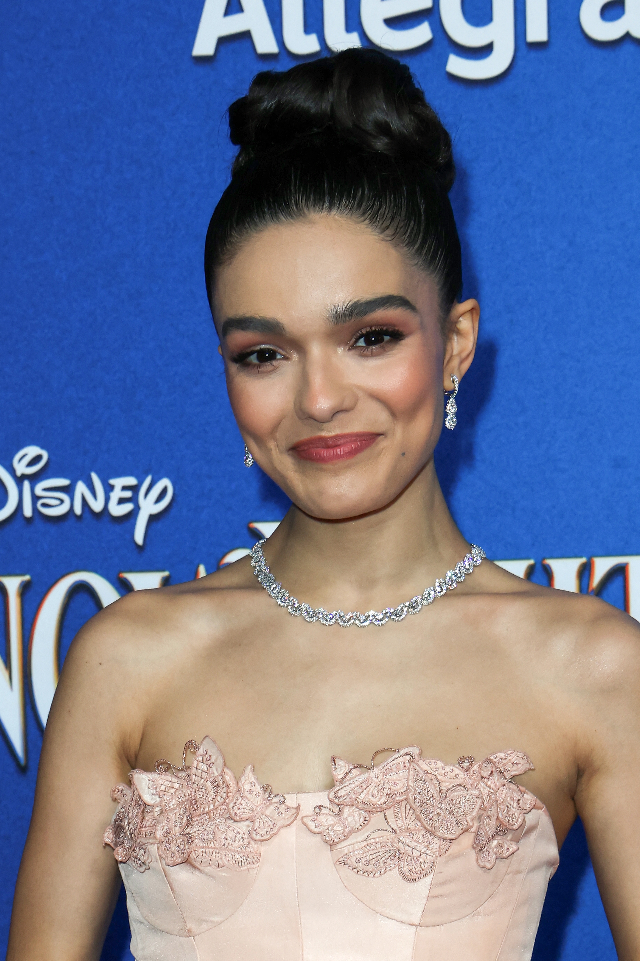 Rachel Zegler at an event, wearing a strapless dress with floral embroidery detail and a necklace, poses for a photo in front of a branded backdrop