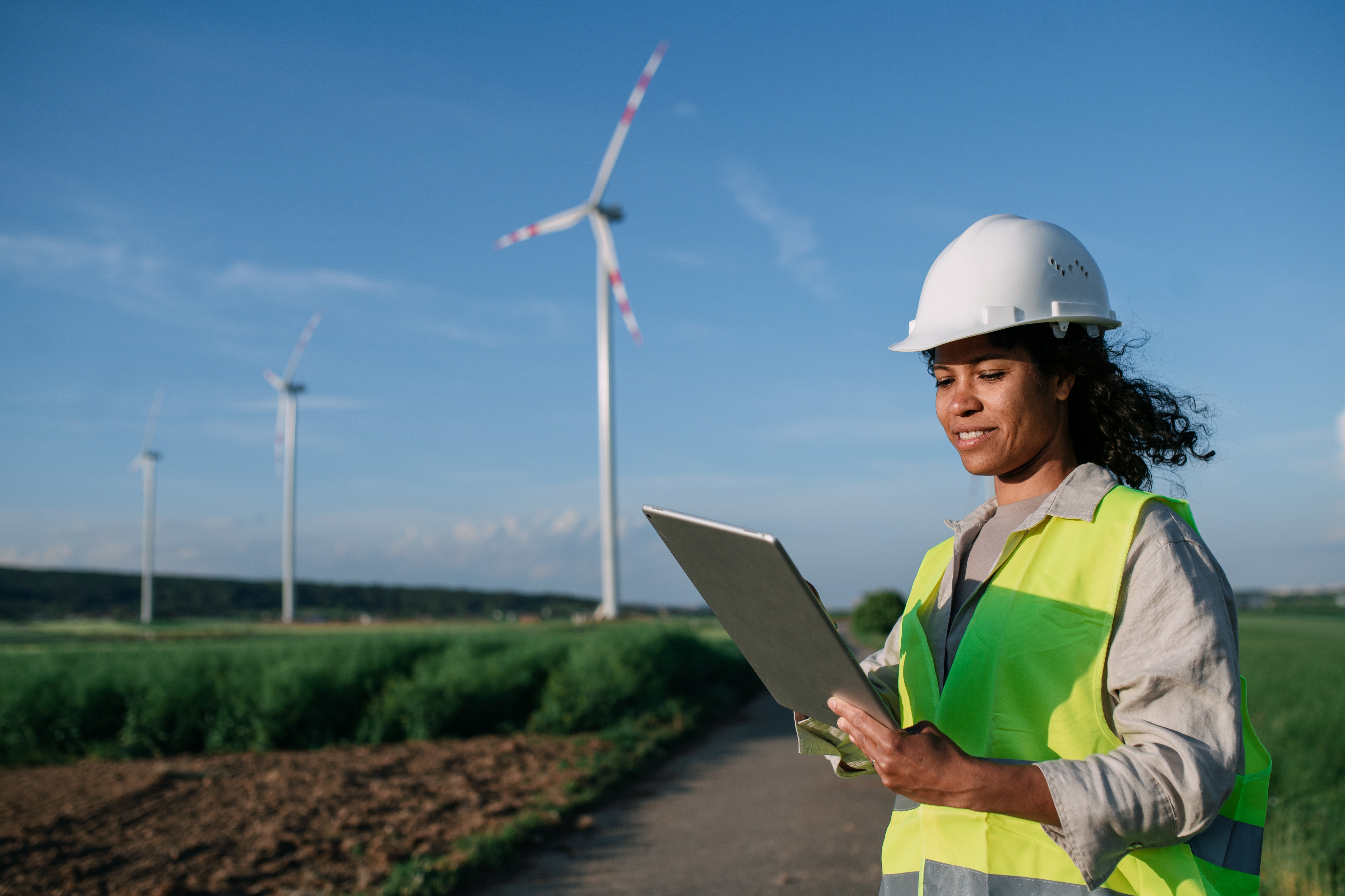 A person in a safety vest and helmet examines a clipboard in a field with wind turbines in the background