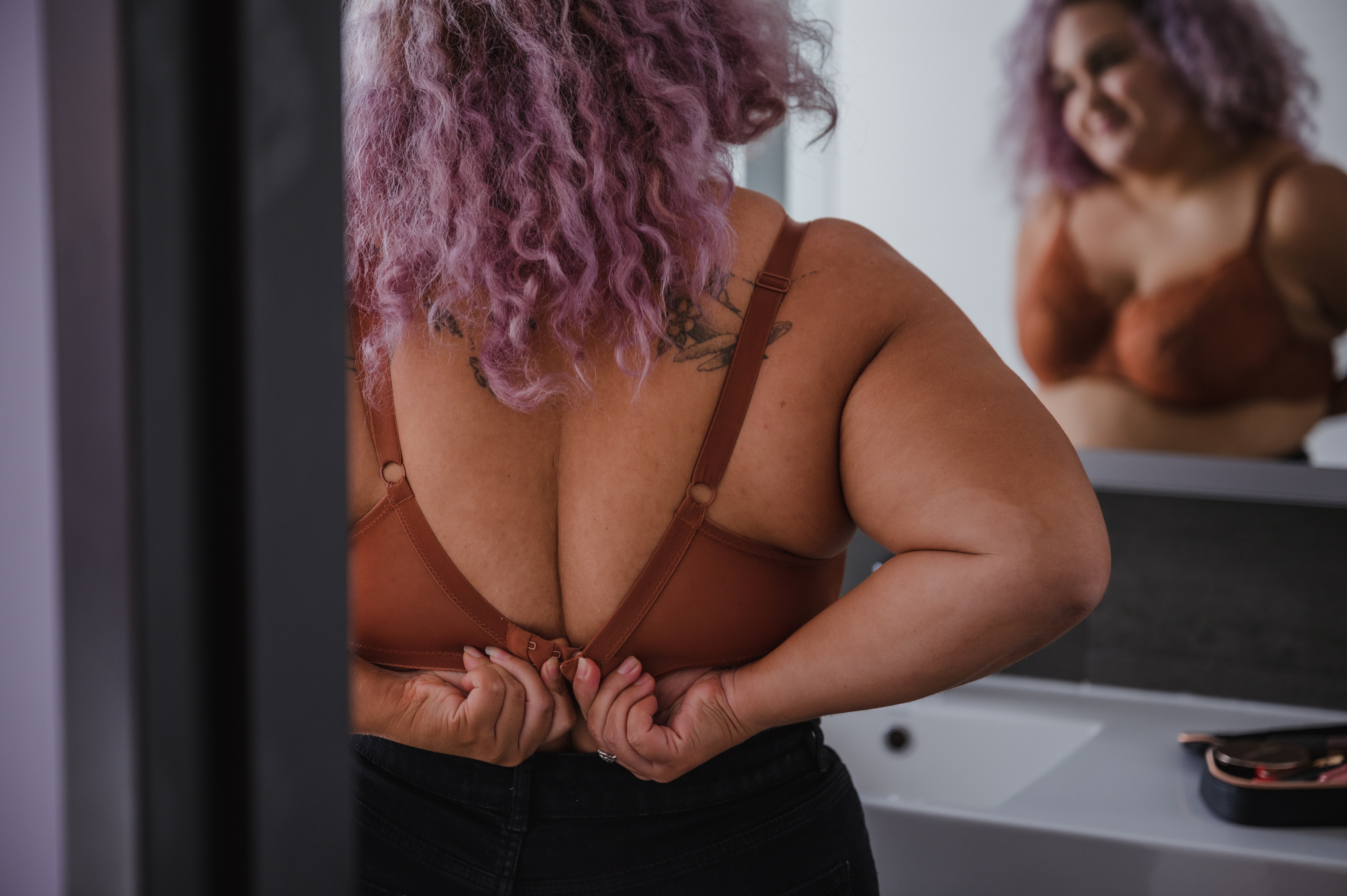 Person with curly hair adjusting their bra strap while looking in a bathroom mirror, showcasing self-reflection or everyday routine